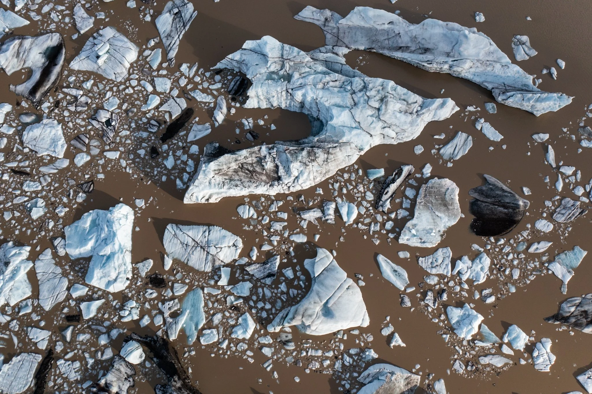 Aerial view of floating icebergs and ice debris in a body of brown water.