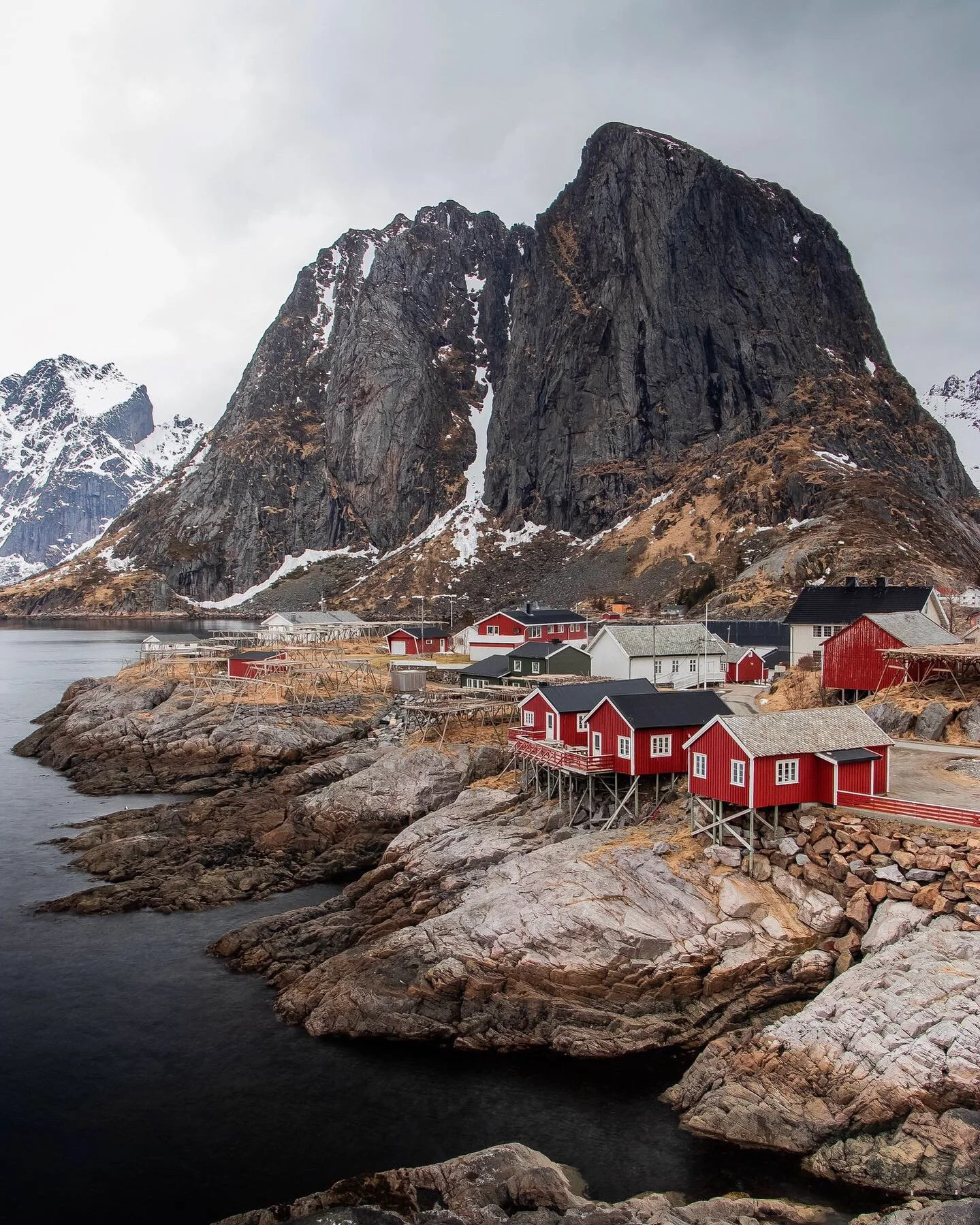 Cliffside village with red and white houses near water, large dark mountain in the background, and overcast sky.