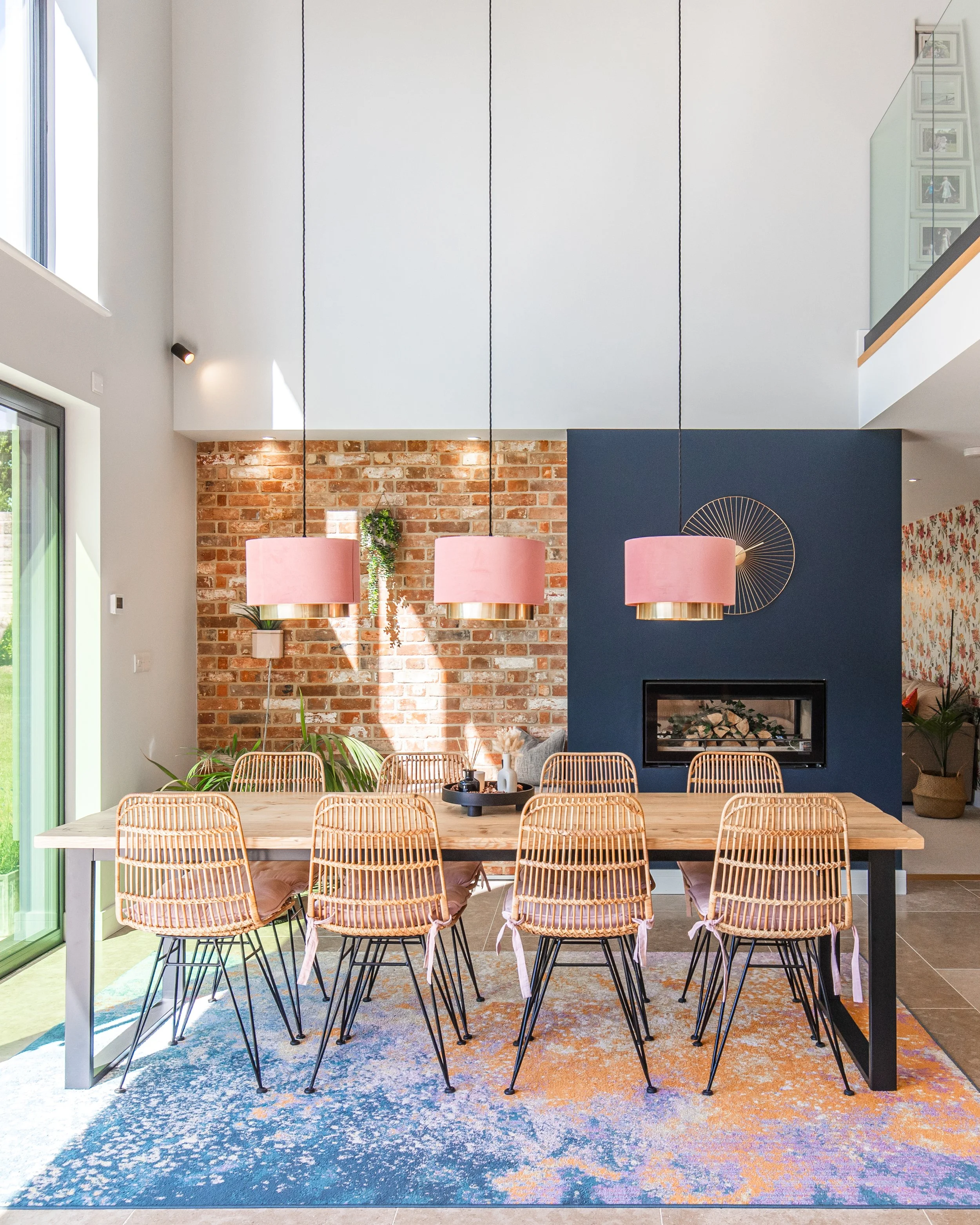 Modern dining room with a large wooden table surrounded by nine wicker chairs, a colorful abstract rug underneath, pink pendant lights hanging above, an exposed brick wall, and a dark blue wall with a decorative metal wall art piece.