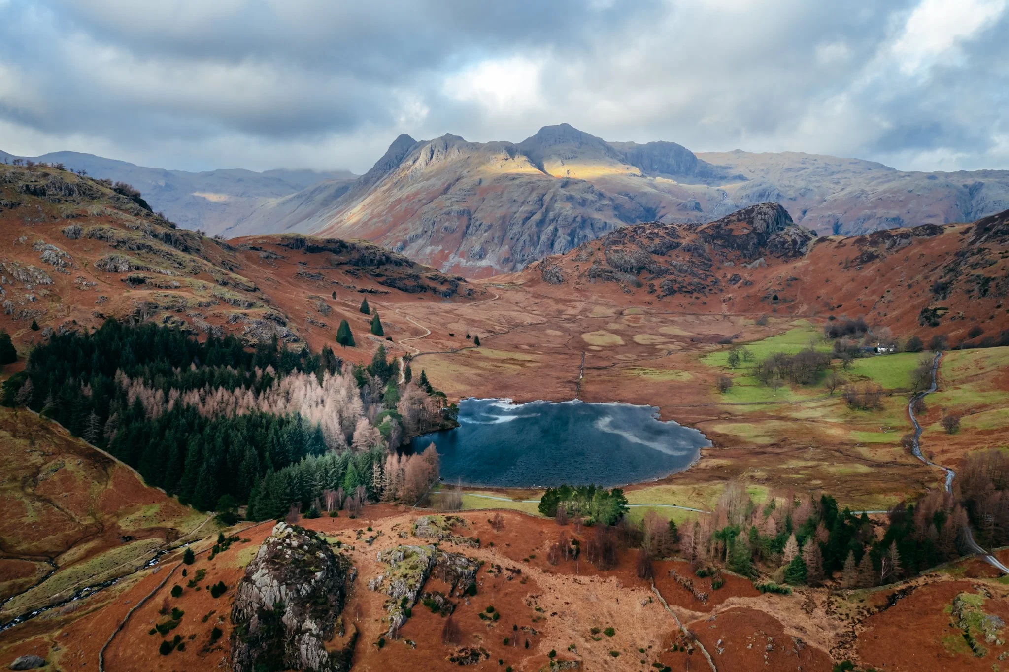 A scenic mountain landscape with rocky peaks, colorful hills, a reflecting lake, and a cloudy sky.