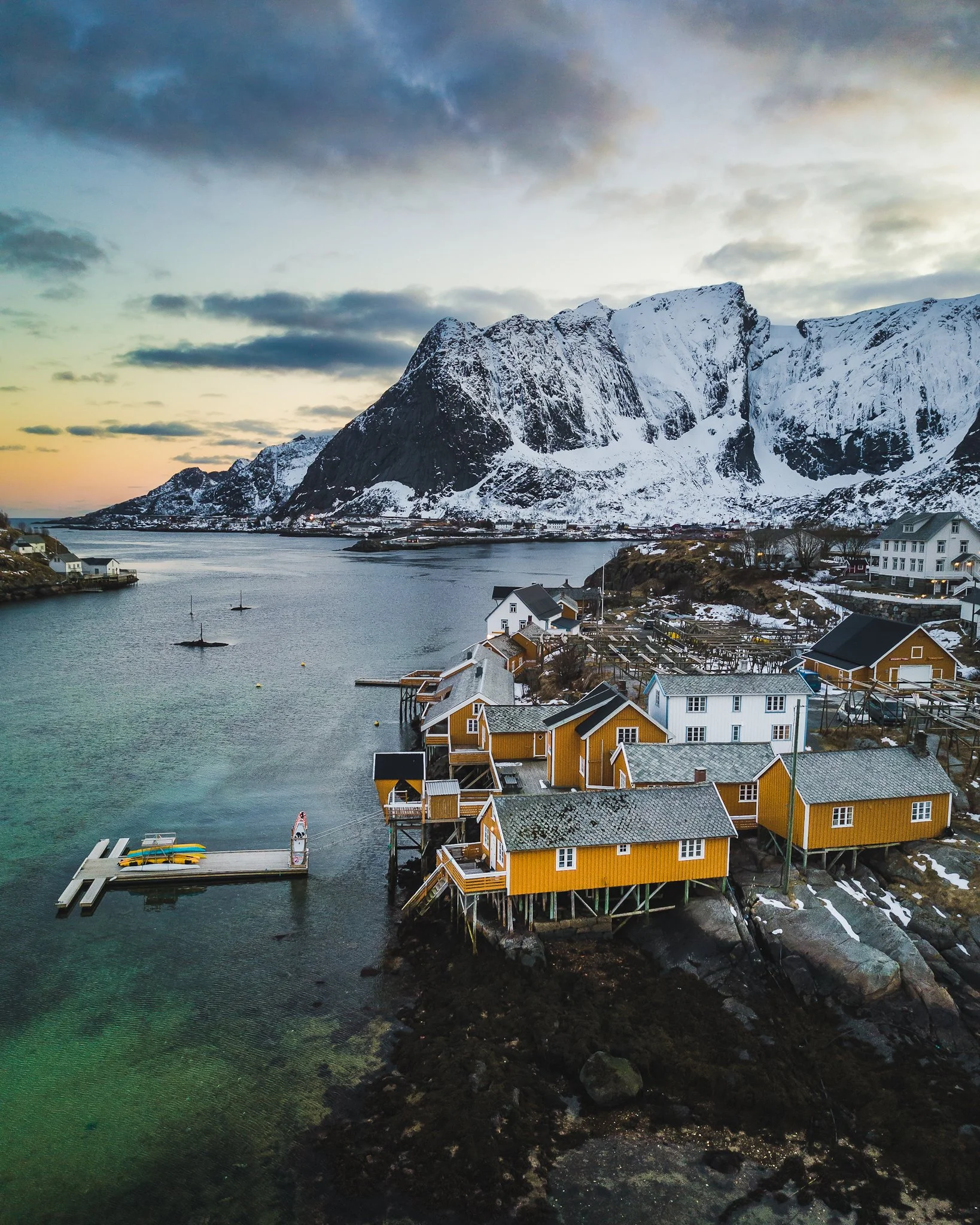 Snow-covered houses along a harbor with a mountainous landscape in the background and cloudy sky.