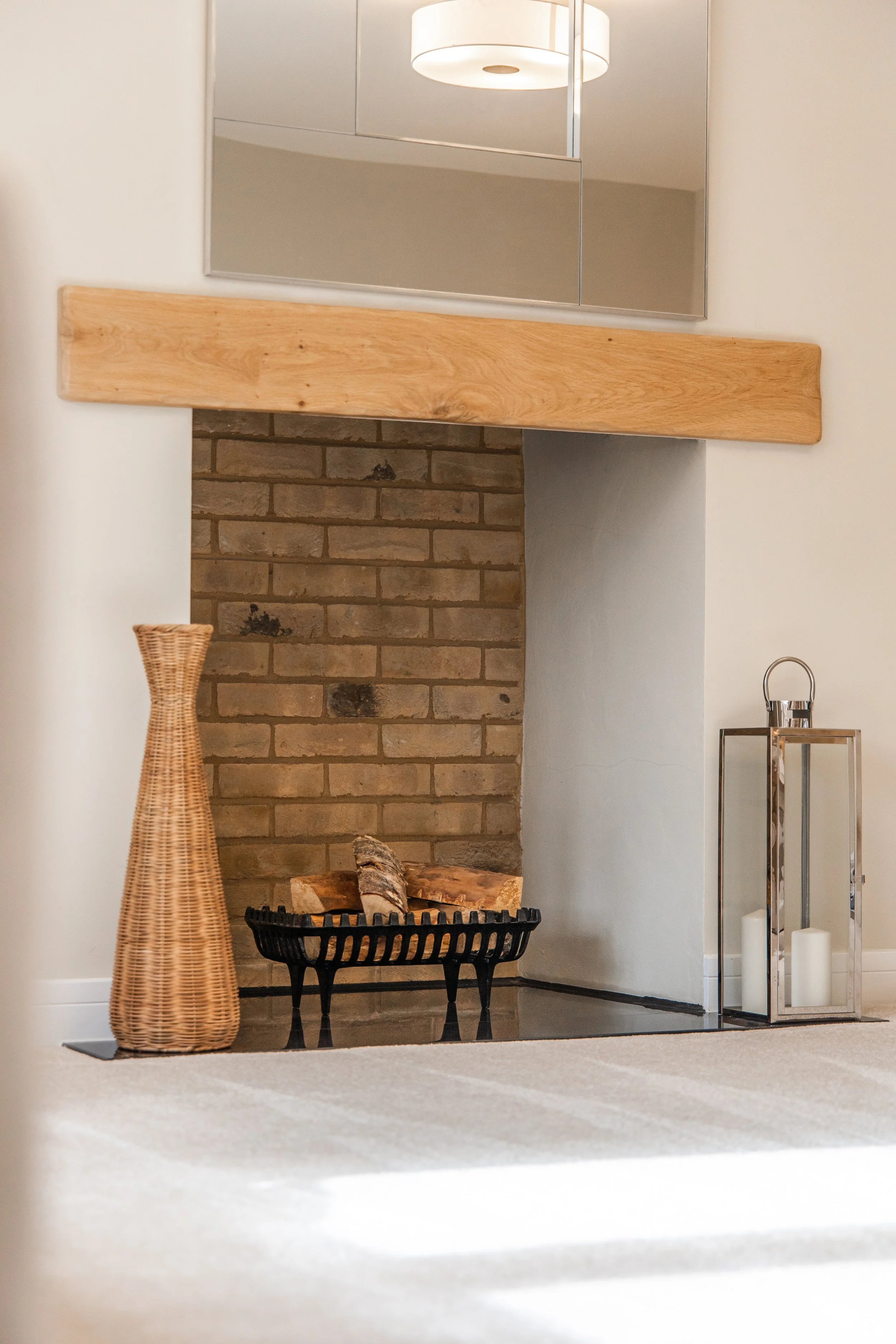 Living room fireplace with a brick hearth, wooden mantel, and a basket of logs, with a wicker vase to the side, and a decorative lantern with candles on the right.