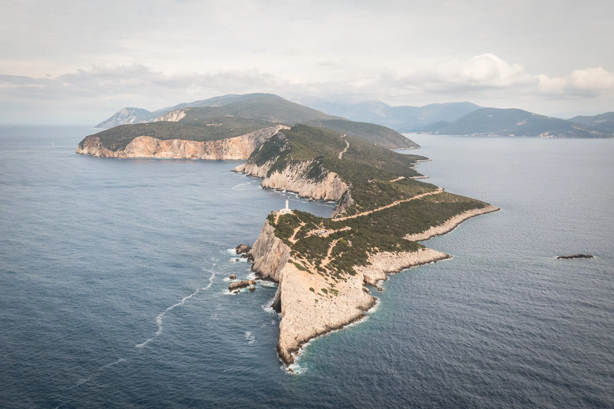 Aerial view of a long, narrow island with rugged, rocky shores covered in greenery, surrounded by the ocean with some waves and a lighthouse at the island's tip, under a cloudy sky.