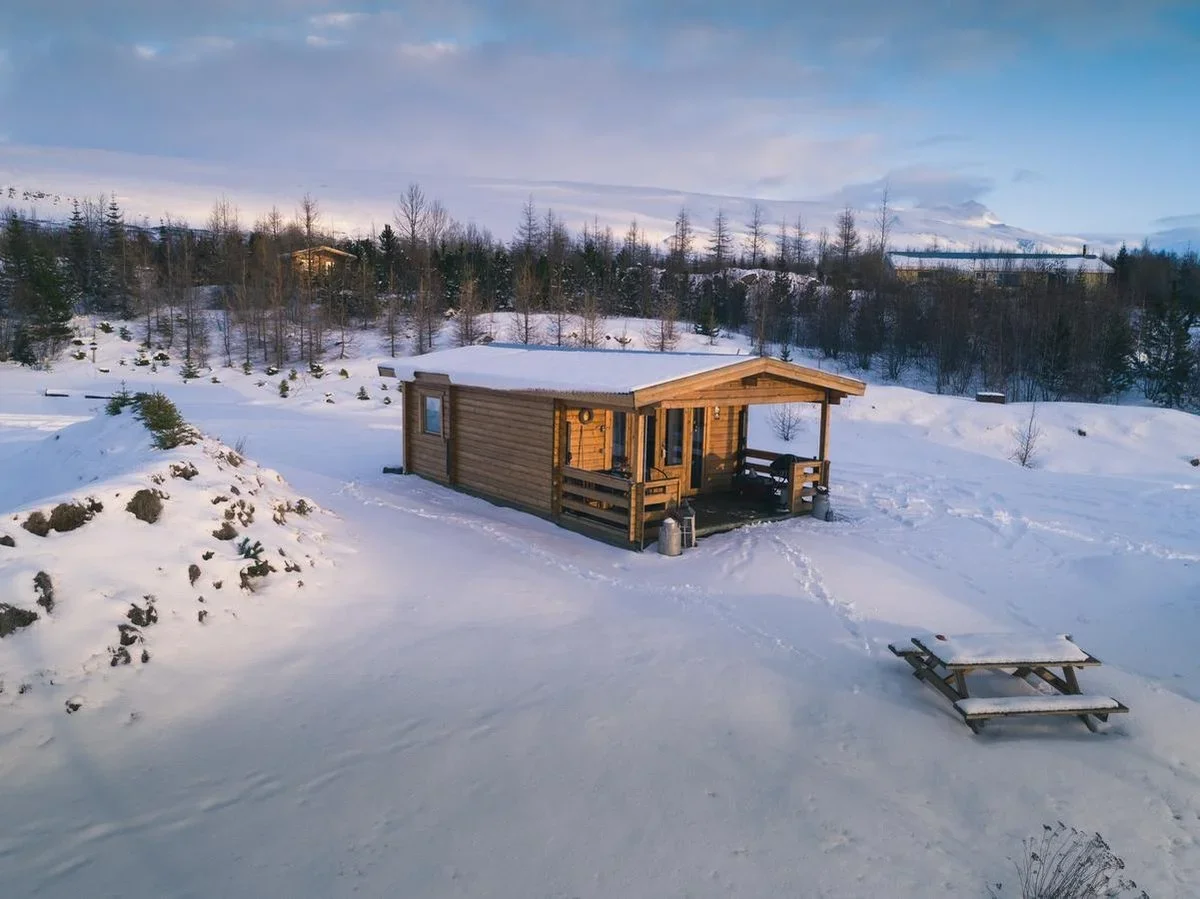 A small wooden cabin in a winter landscape, covered in snow, with a porch and a picnic table nearby, surrounded by trees and snow-covered hills.