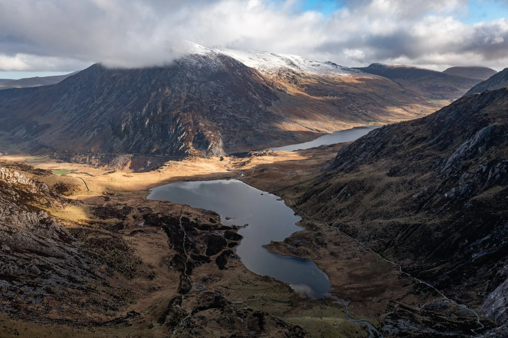 Mountain landscape with snow-capped peaks, clouds, lakes, and rugged terrain in a valley.