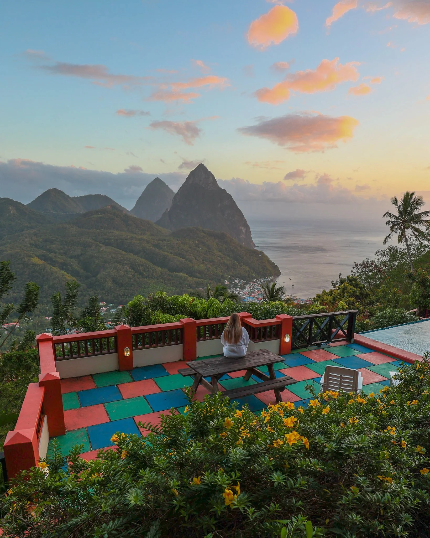 A scenic view of mountains, the ocean, and a colorful terrace with a person sitting and enjoying the view at sunset.