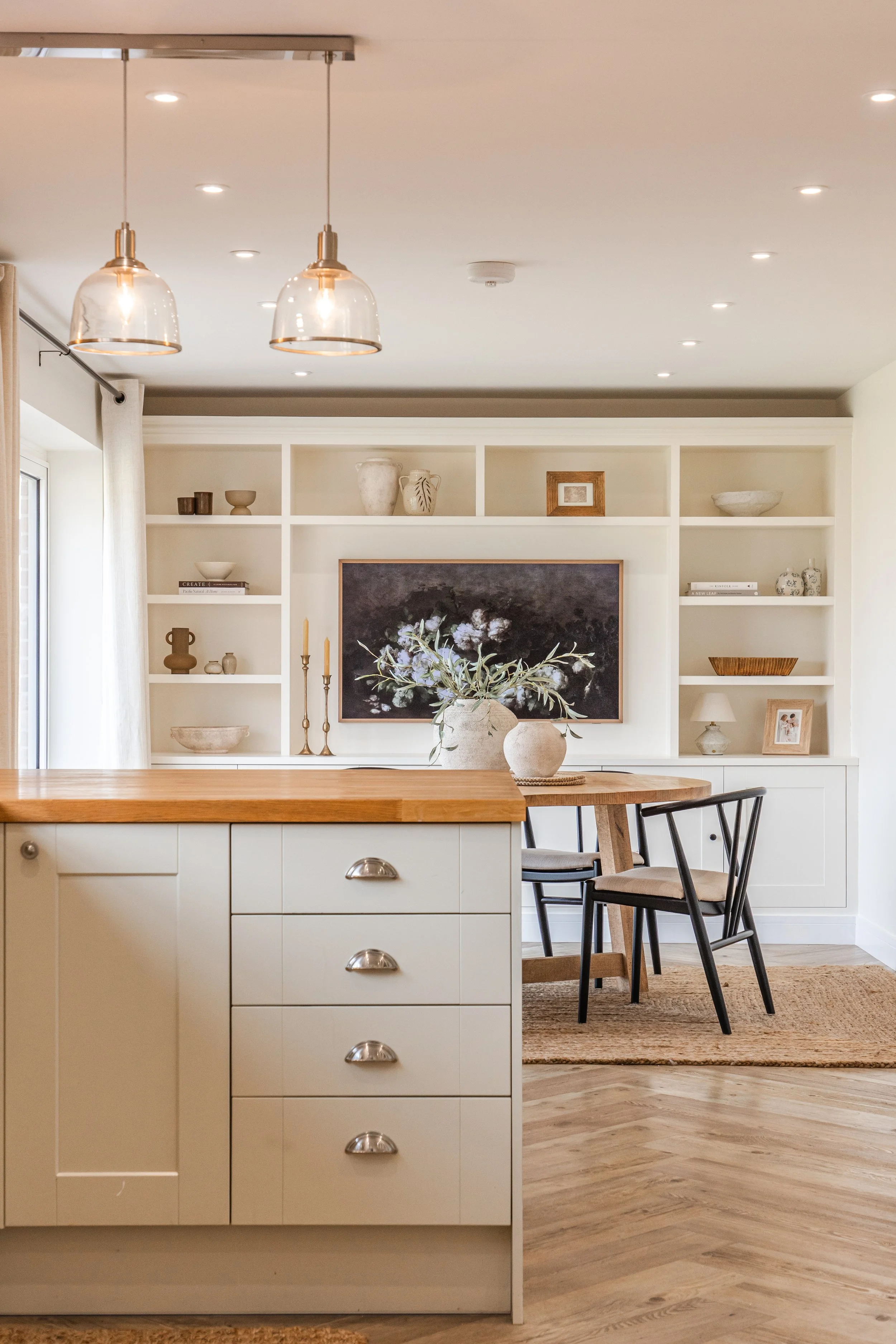 A kitchen with a white island and beige cabinetry, decorated with vases and candles, overlooking a dining area with black chairs, a wooden table, and built-in shelves with decorative items and books, and a large hanging pendant light.