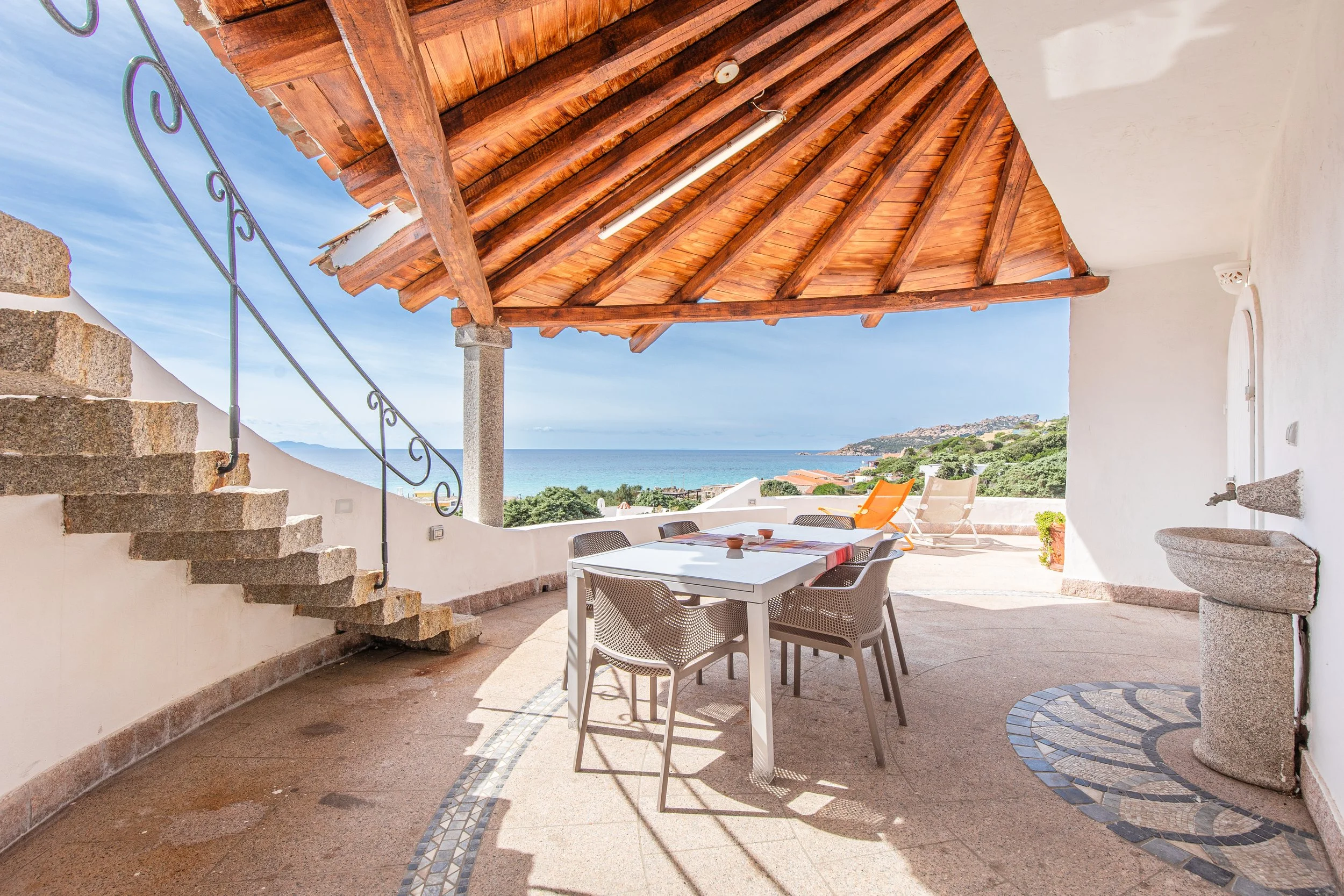 Outdoor terrace with a white table and four chairs, overlooking the ocean, with a wooden roof and white walls, a staircase on the left, and lounge chairs in the background.