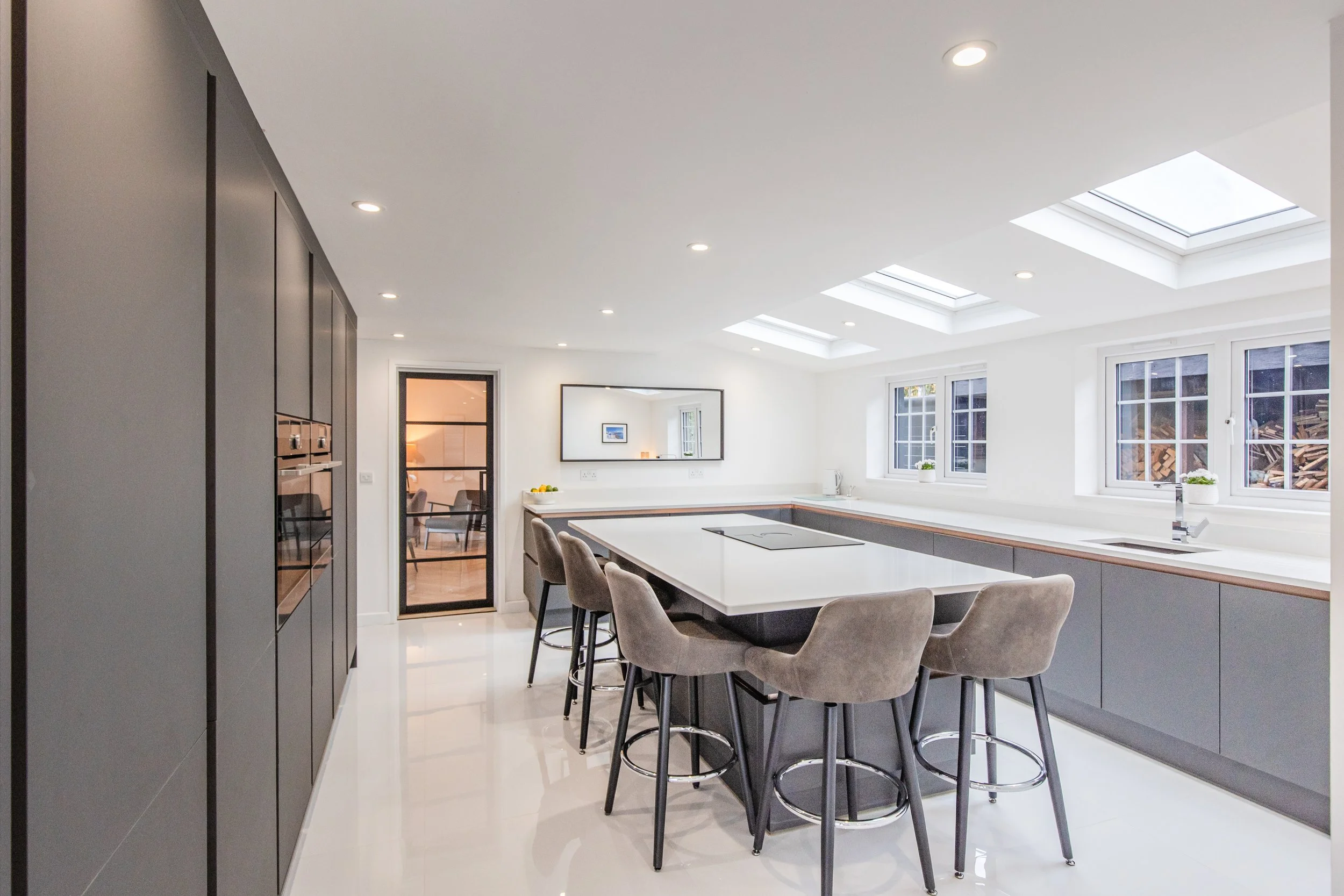 Bright modern kitchen with white counters, gray cabinets, beige bar stools, and skylights in the ceiling.