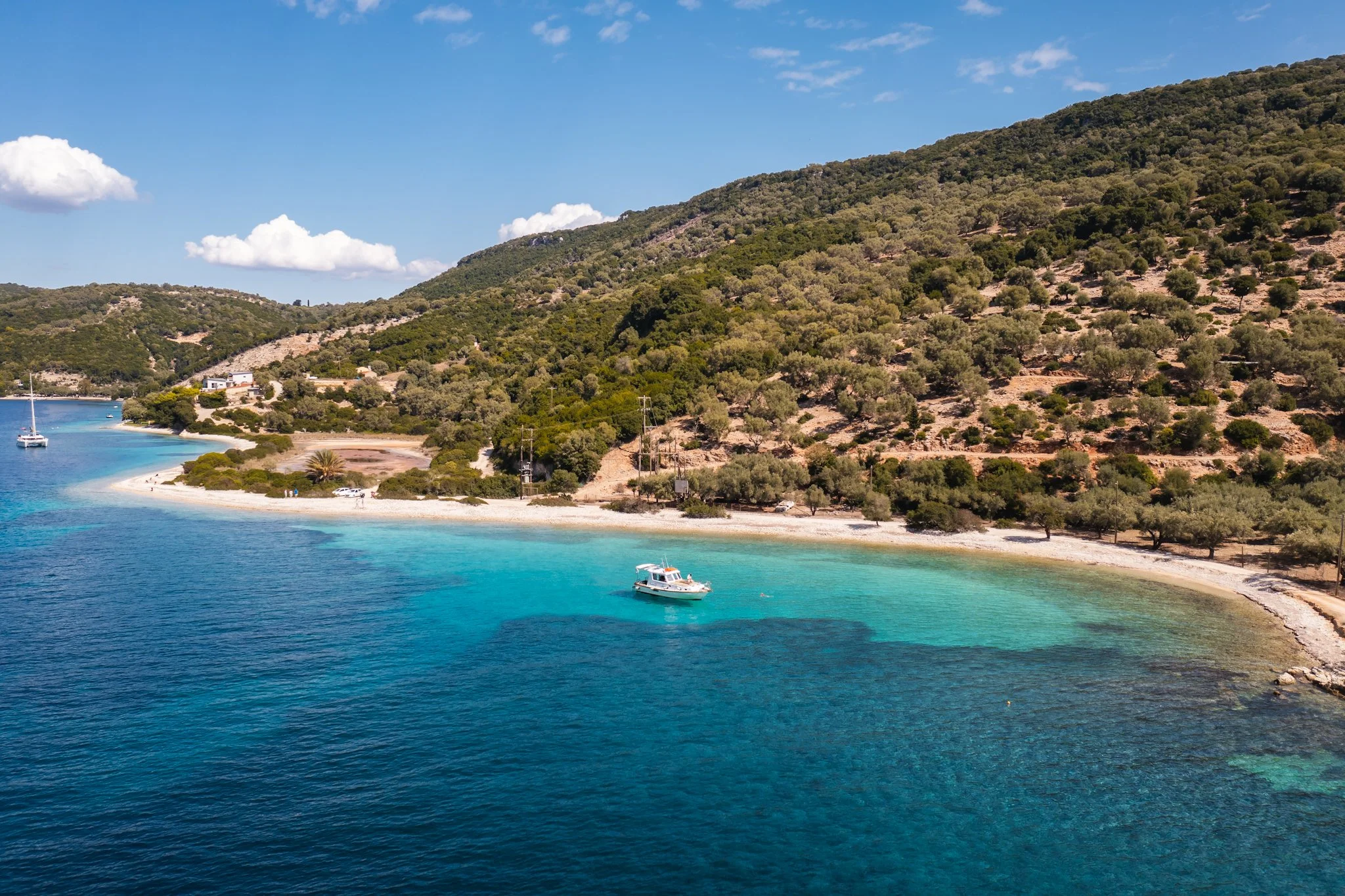 Scenic view of a bay with turquoise water, a sailboat, and a motorboat near the shore, with green forested hills and a blue sky with clouds in the background.
