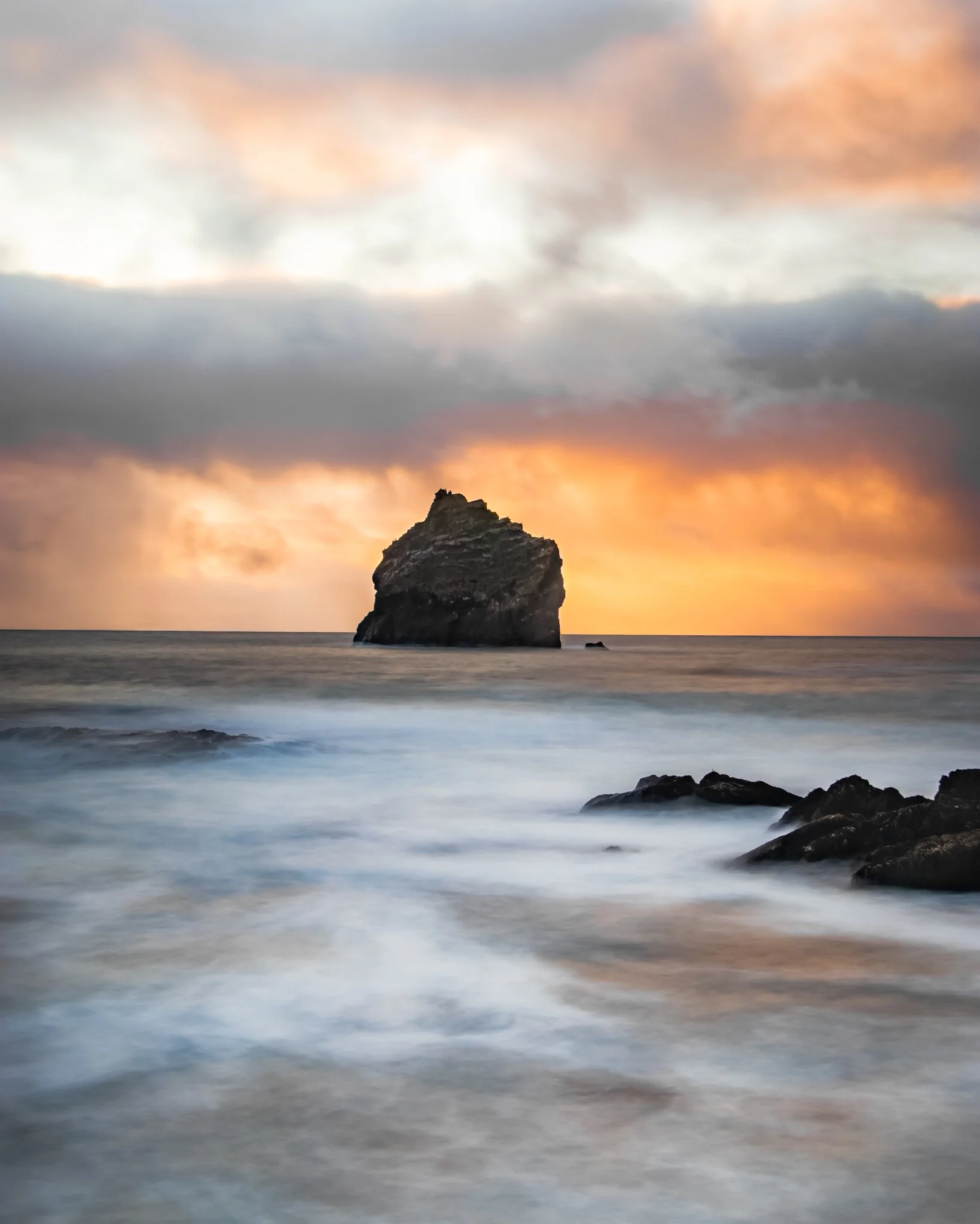 A large rock formation rises from the water against a sunset sky with orange and pink clouds.