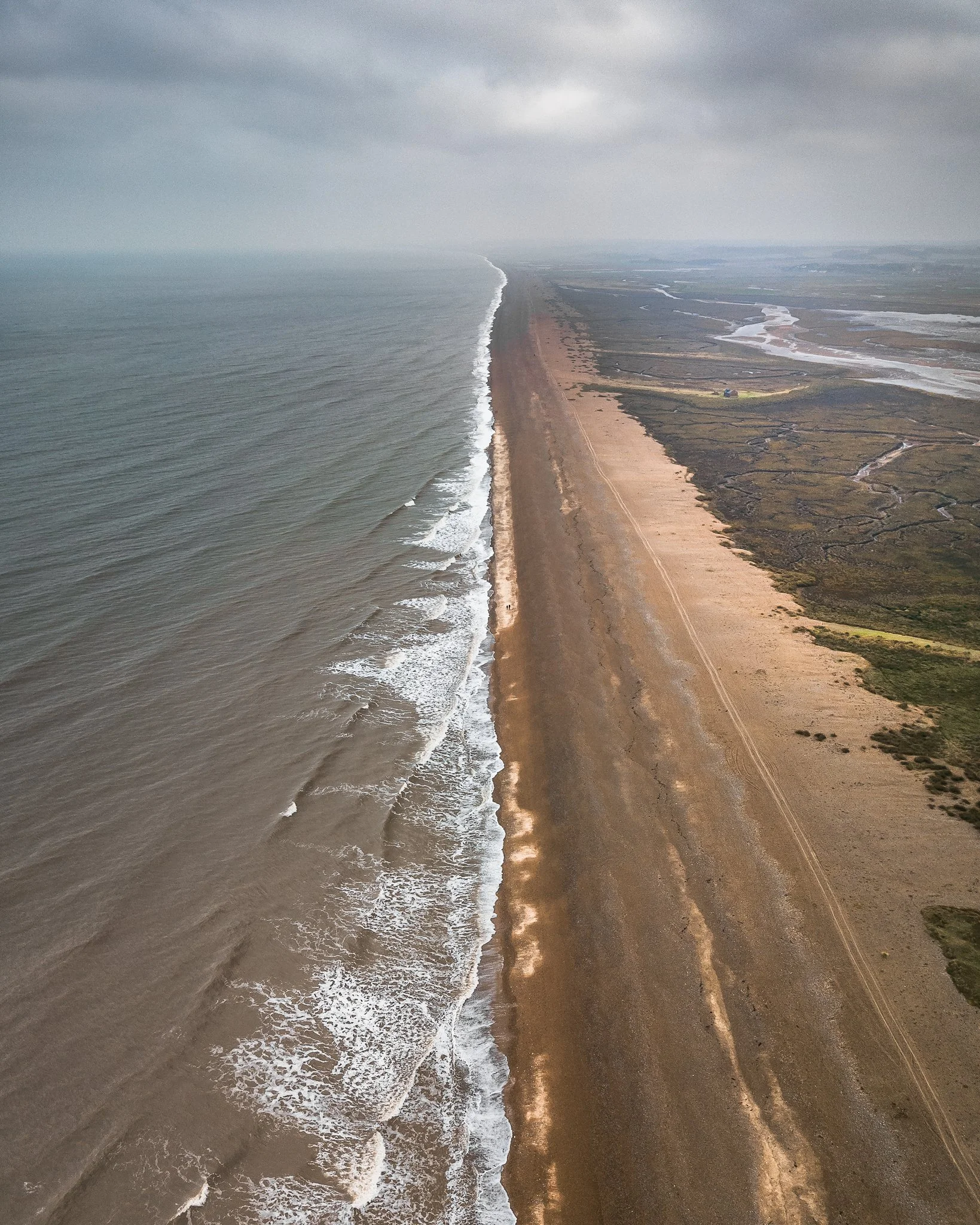 Aerial view of a long sandy beach with waves crashing onto the shore, backed by marshland and a cloudy sky.