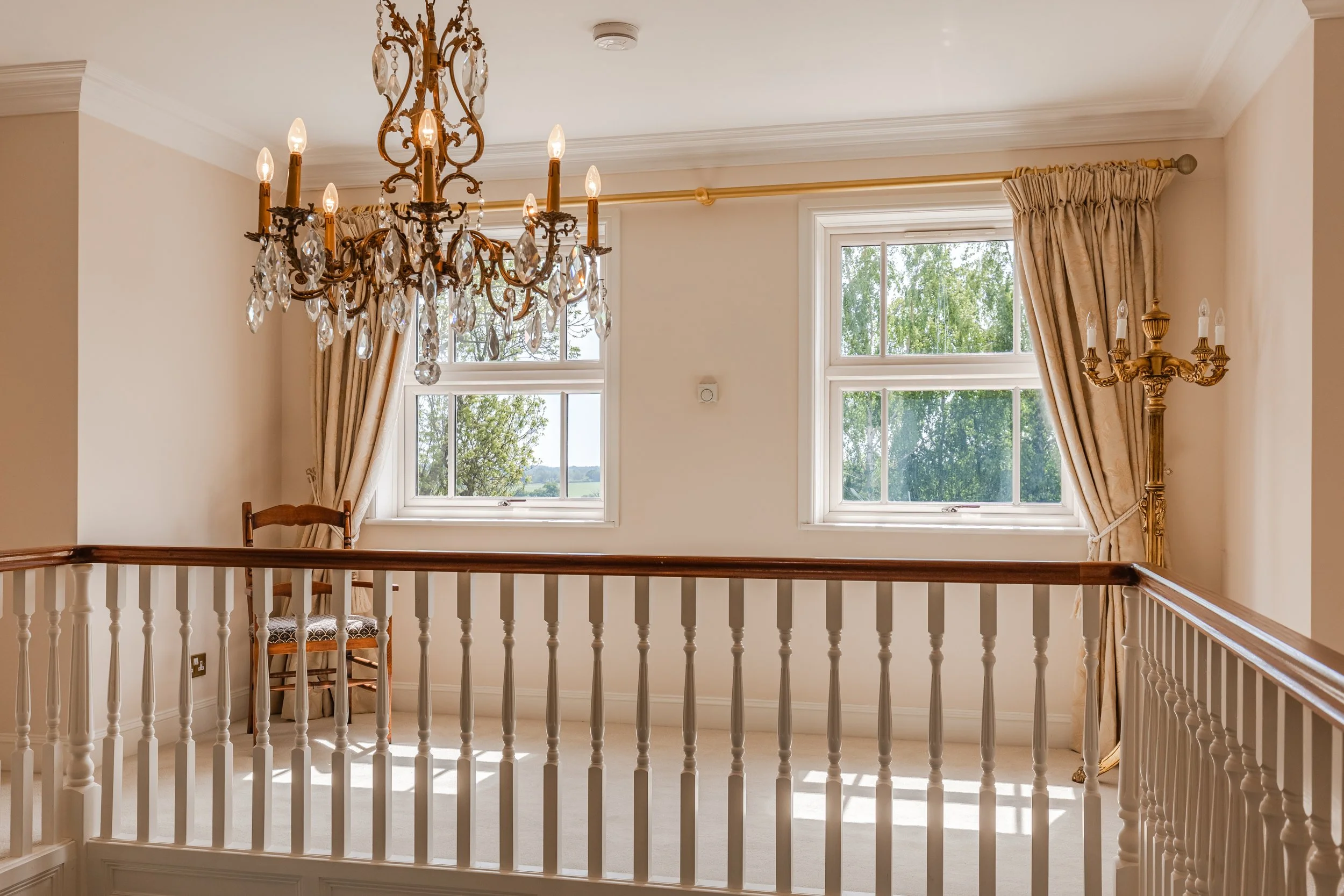 Interior view of a bright living space with large windows, beige curtains, a chandelier, a candelabra, and a white railing.
