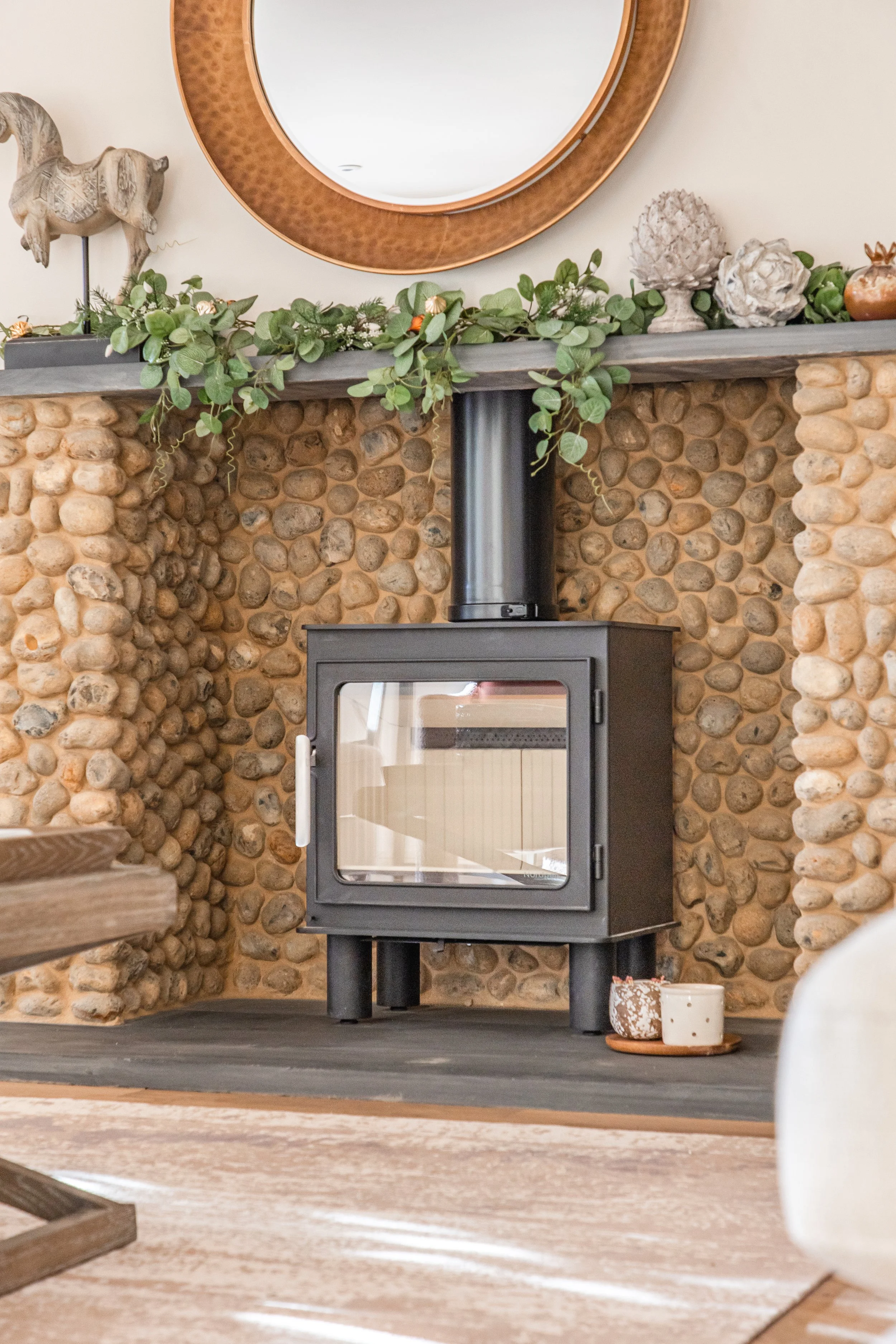 A cozy living room fireplace with a wood stove set against a wall with rounded river rocks. Decorative objects including a carved elephant, pinecone sculptures, and potted greenery decorate the mantel. A large round mirror with a copper frame hangs a