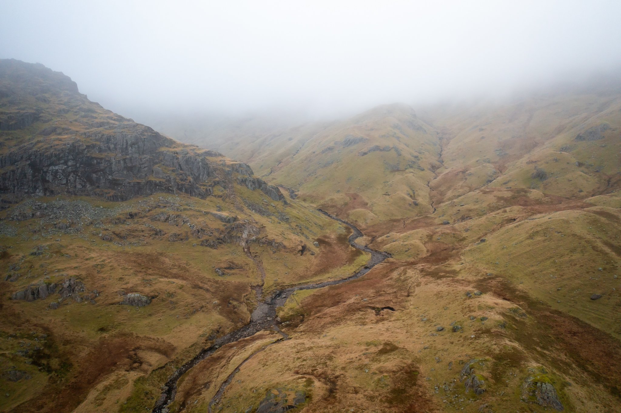 Mountain landscape with rocky slopes and a winding stream under a foggy sky.