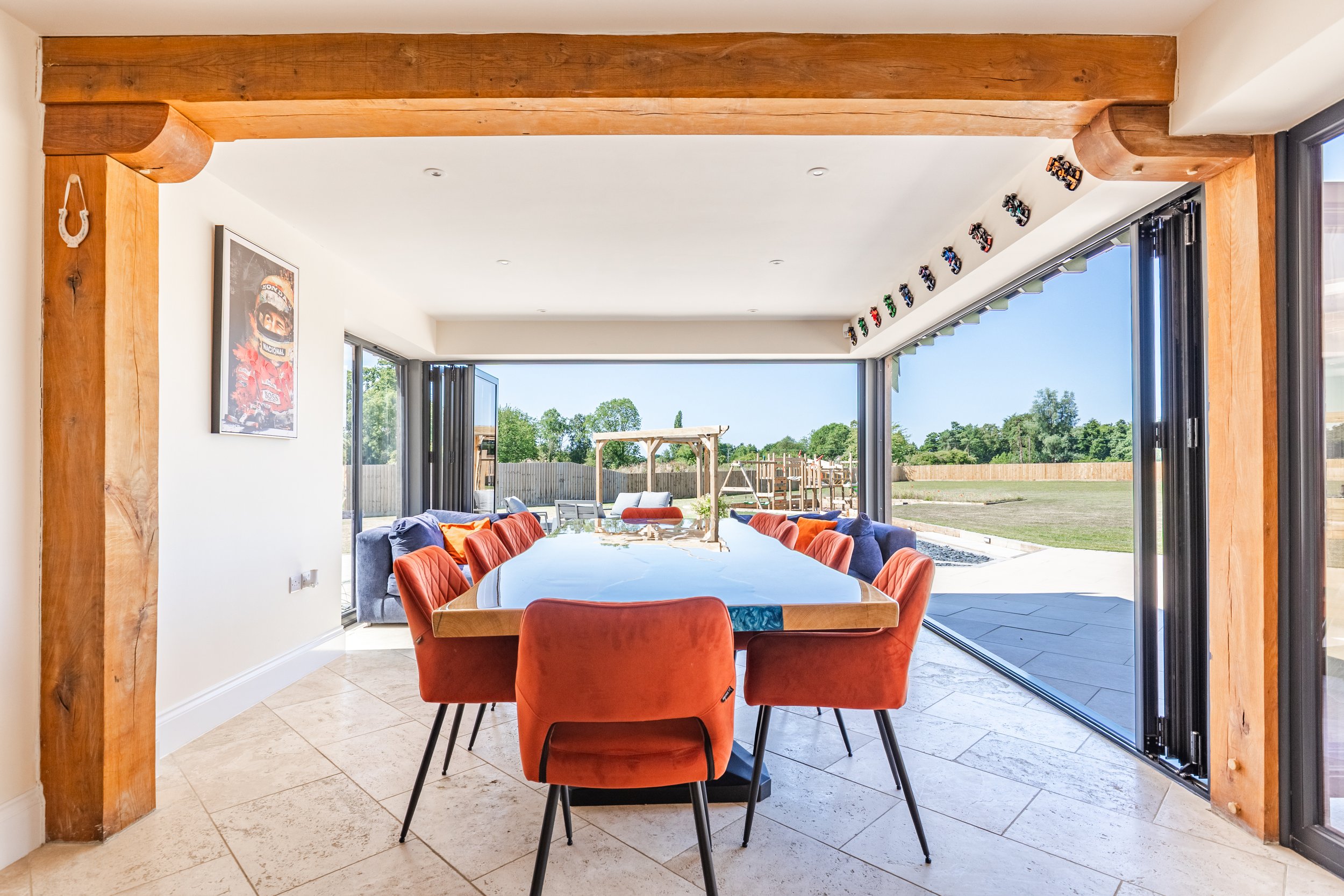 A dining room with a large wooden table surrounded by orange and gray chairs, with a view of a backyard with a wooden playset and green trees outside through sliding glass doors. The room has a wooden beam above and a picture of a racing driver on th