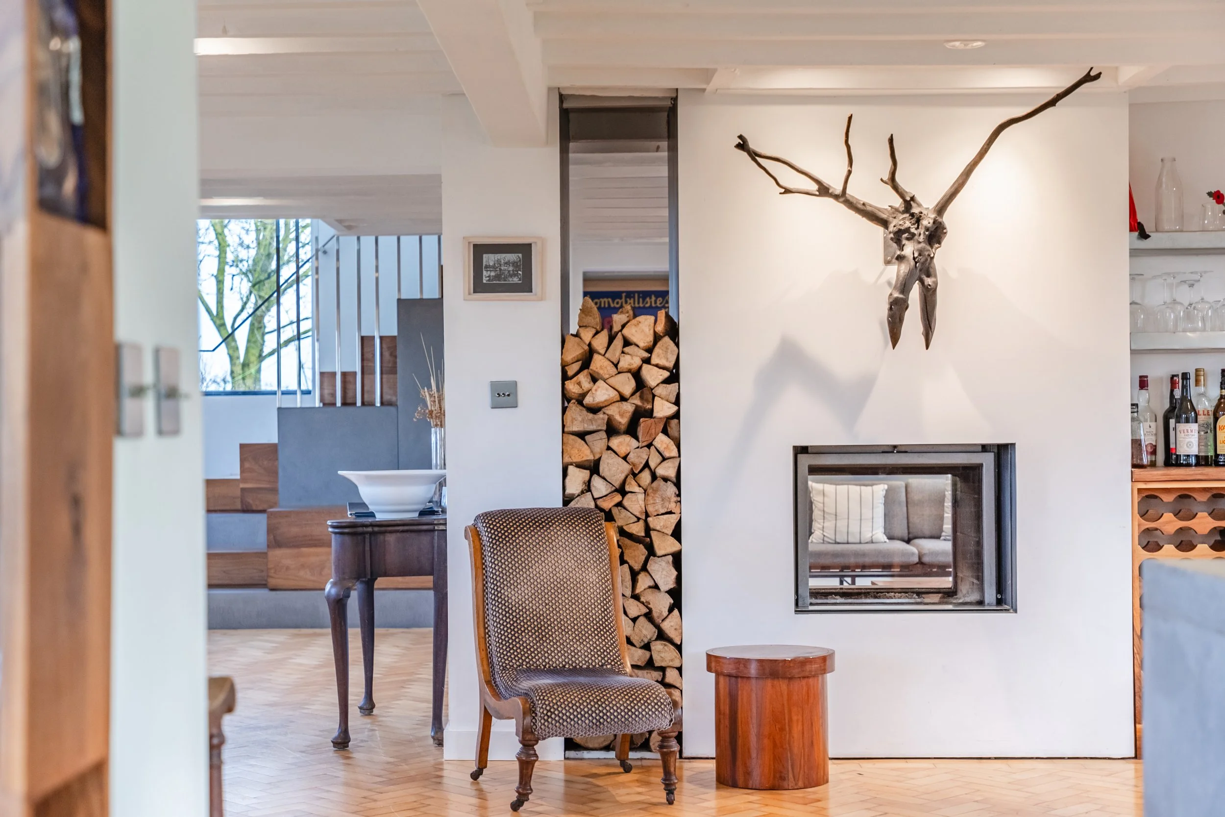 Living room with wood pile stacked vertically beside a fireplace, a wooden chair with a patterned cushion, a small side table, decorative items, and large windows showing trees outside.