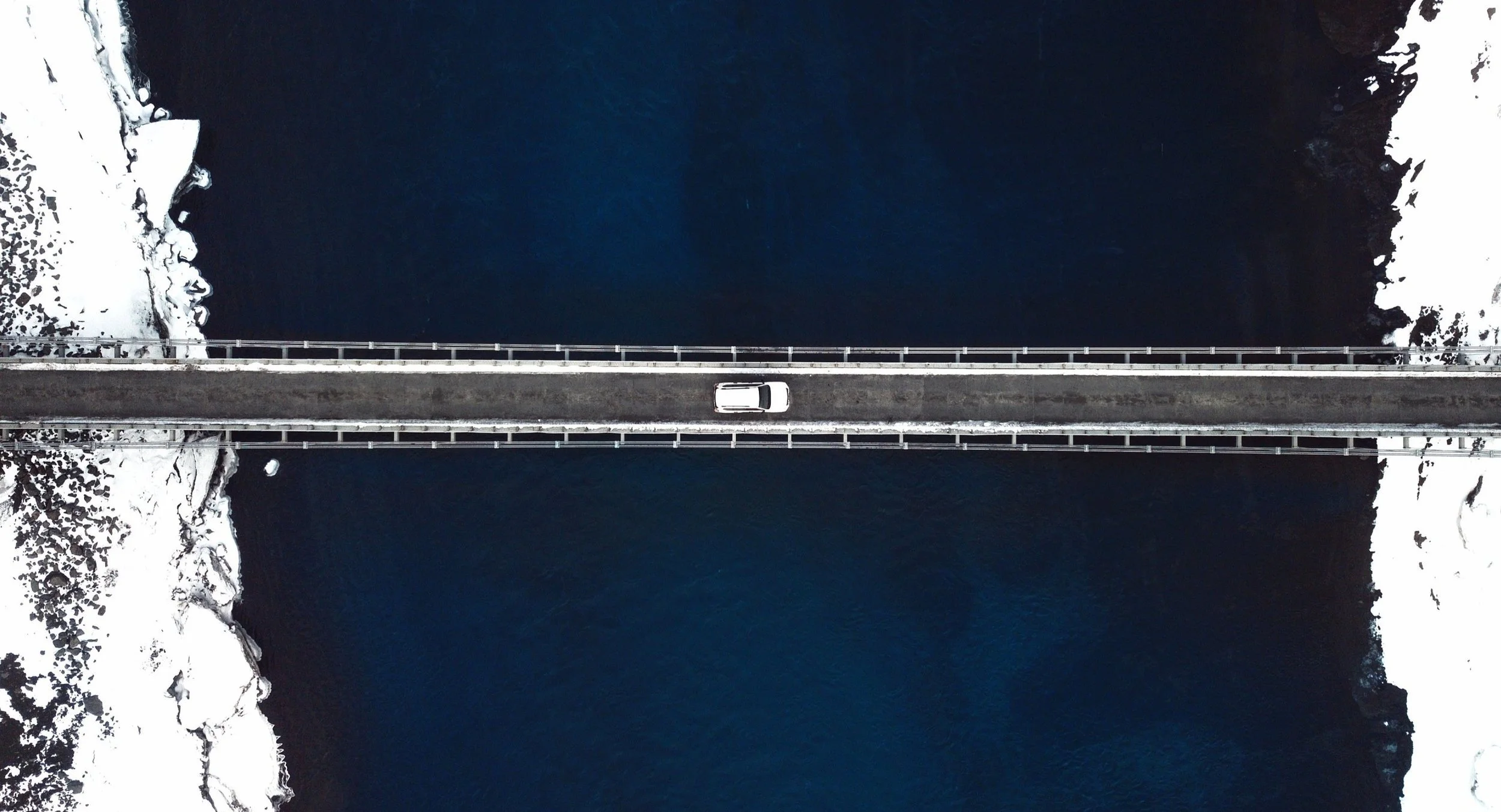 An aerial view of a bridge crossing a body of dark blue water with snow-covered land on both sides.