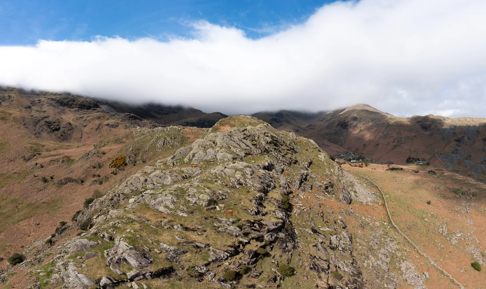 A scenic mountain landscape with rocky terrain in the foreground, rolling hills in the middle, and mountains partly covered by low-hanging clouds under a partly cloudy blue sky.