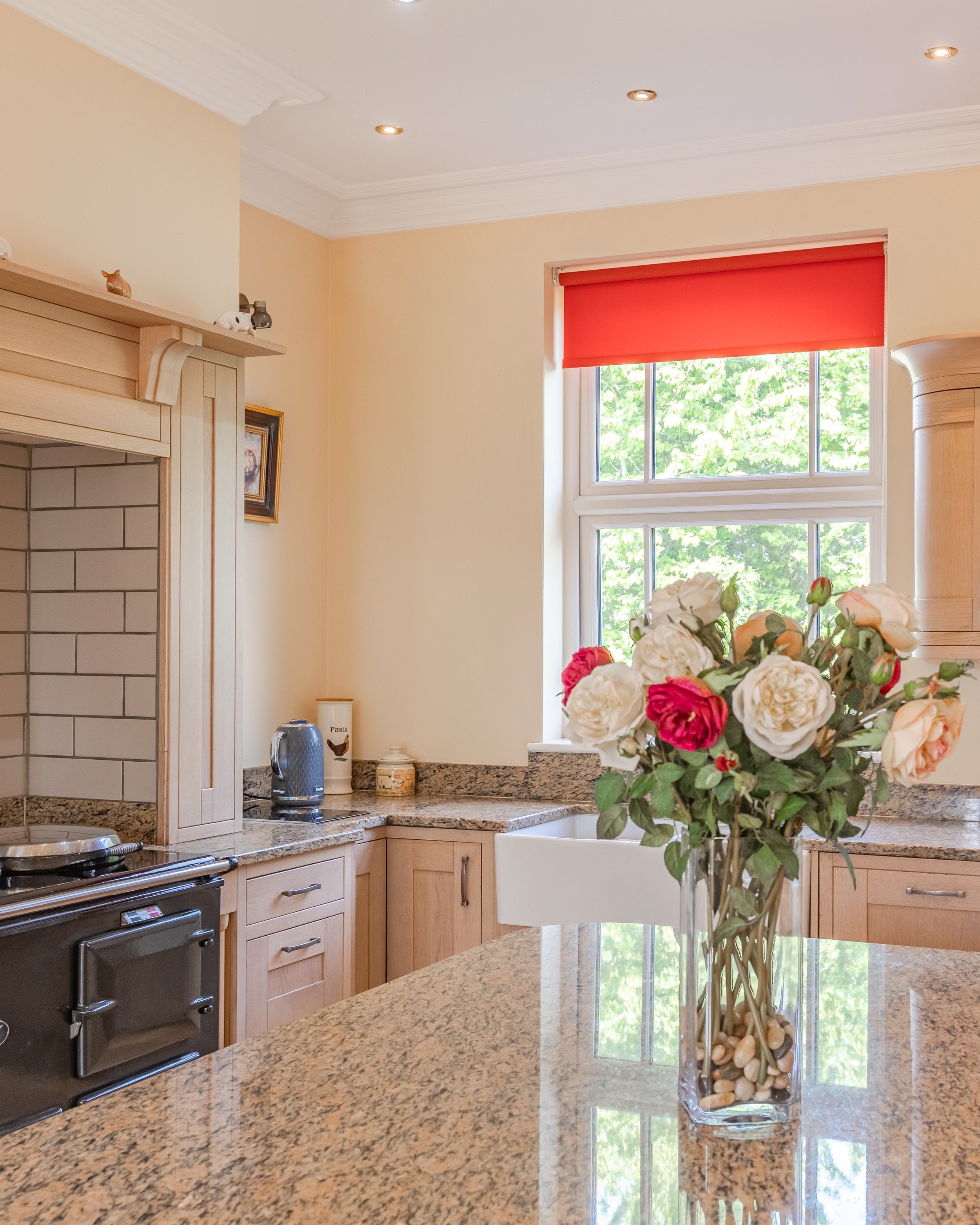 A kitchen with granite countertops, light wooden cabinets, a window with a red valance, a bouquet of roses in a glass vase, and small decorative items on the wall and counters.