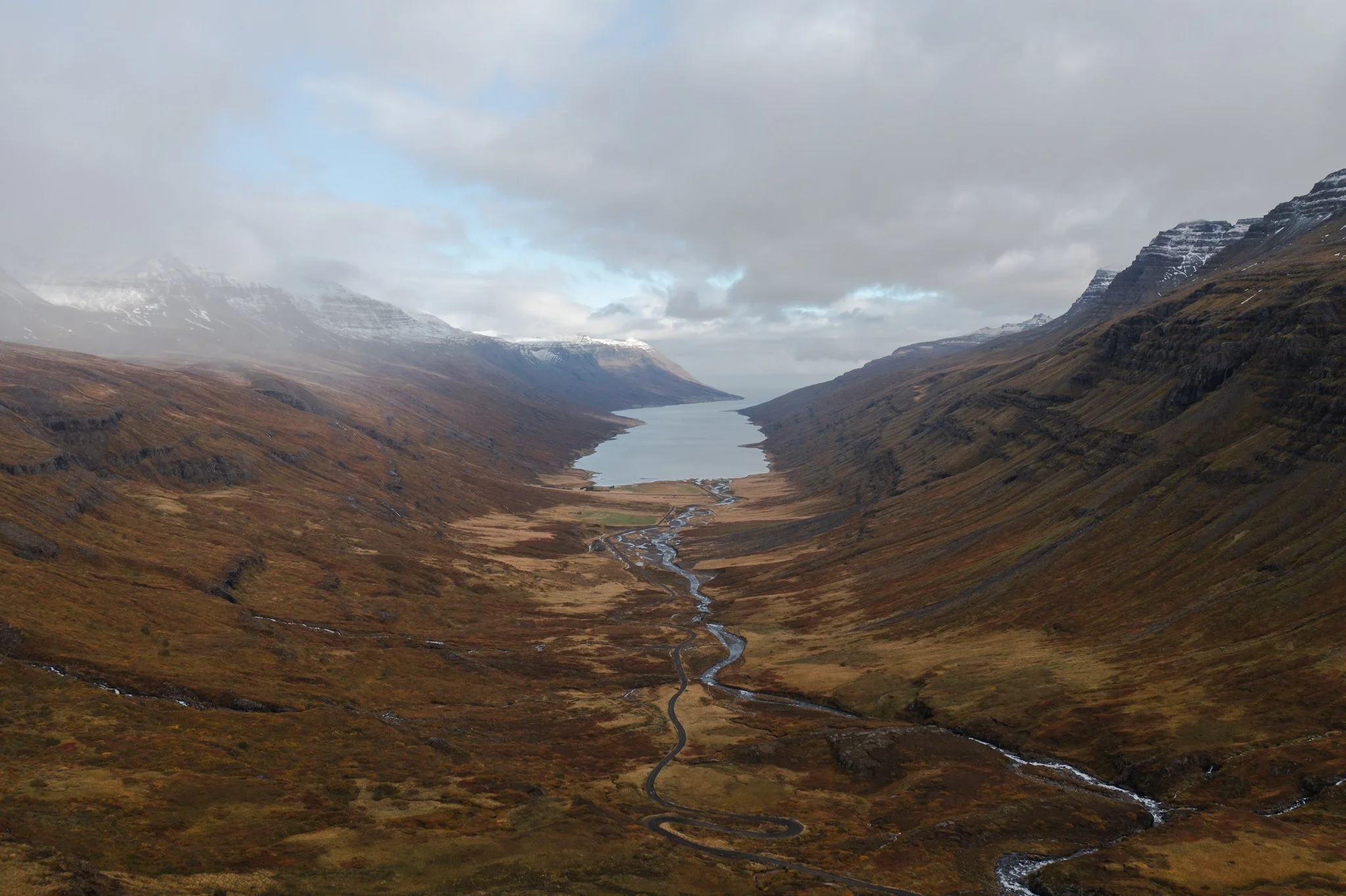 A wide view of a mountain valley with a winding river, snow-capped peaks in the background, and cloudy sky overhead.