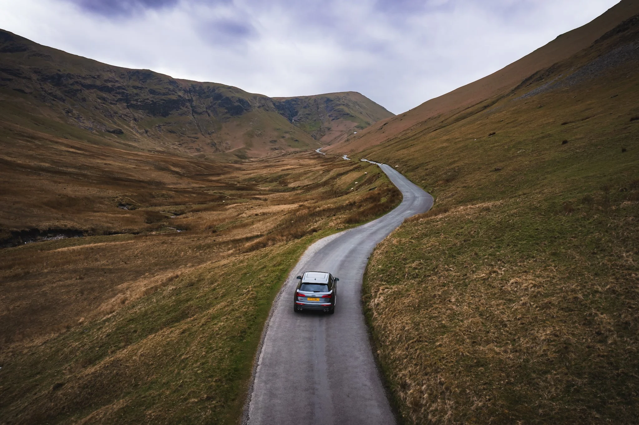 A winding road running through a hilly, grassy landscape with mountains in the background and a car driving on the road.