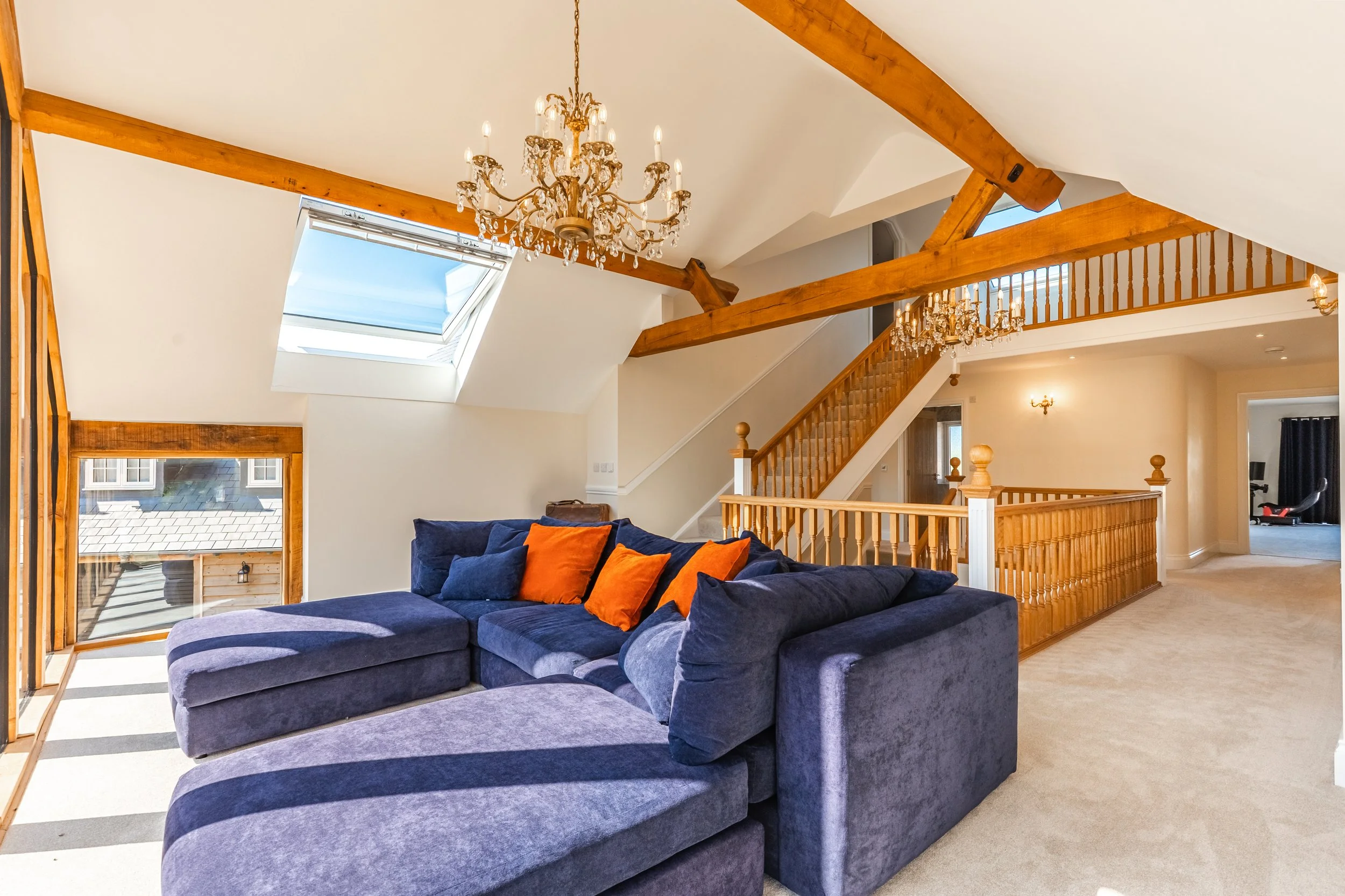 Living room with a large blue sectional sofa with orange and blue pillows, wooden beams and chandelier, skylight window, and staircase leading to upper level.