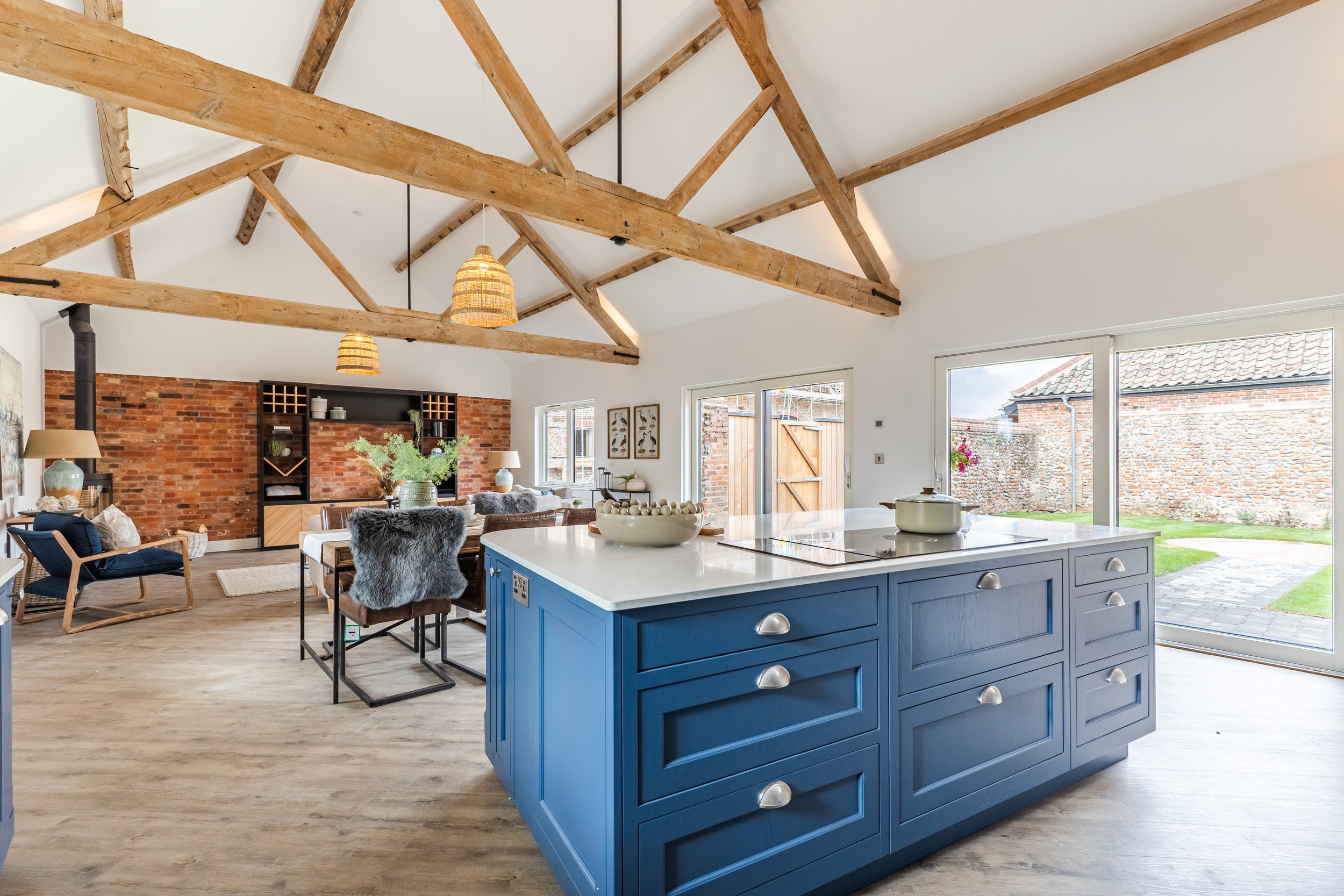 Open-concept living room and kitchen with exposed wooden beams, brick accent wall, large windows, and a central blue kitchen island.
