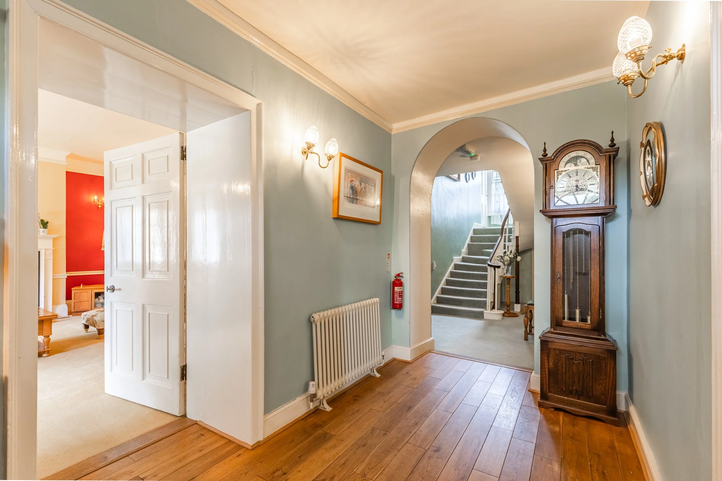 A hallway with light green walls and wooden floorboards, featuring a white radiator, a grandfather clock, framed pictures, wall sconces, a staircase with carpeted steps, and a doorway leading to a room with red walls and white trim.
