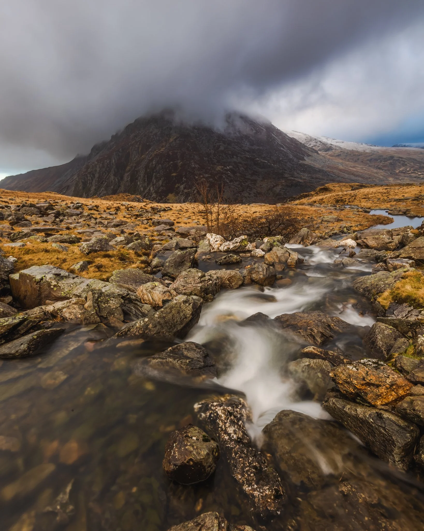 A mountain with a rocky surface and a glacier on the top right, partially covered by dark, heavy clouds. In the foreground, a flowing stream with visible rocks and patches of grass on the banks.