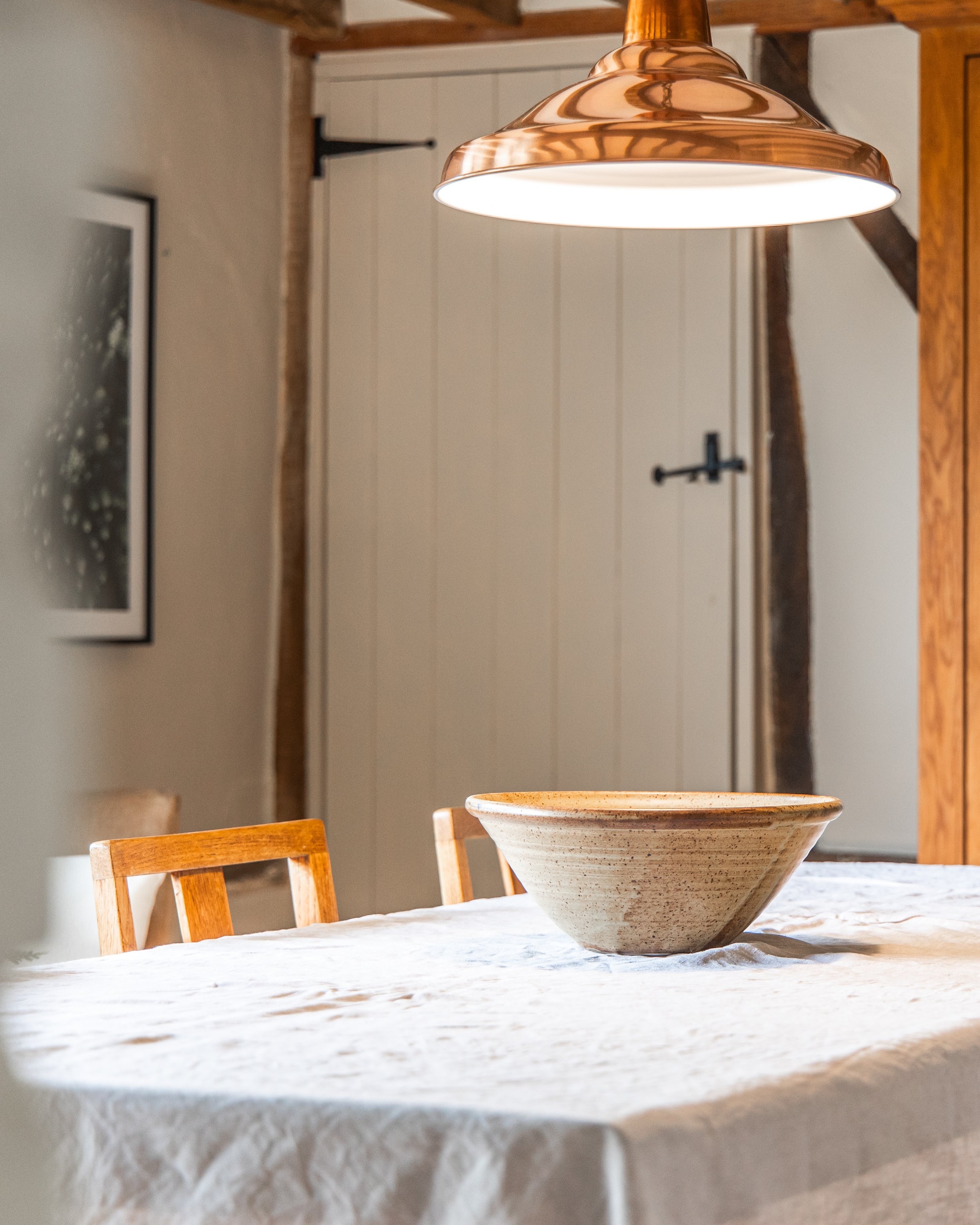 Rustic dining room table with a beige, textured tablecloth, a large ceramic bowl, wooden chairs, and a copper pendant light hanging above. There is a framed picture on the wall and a door with black hardware in the background.