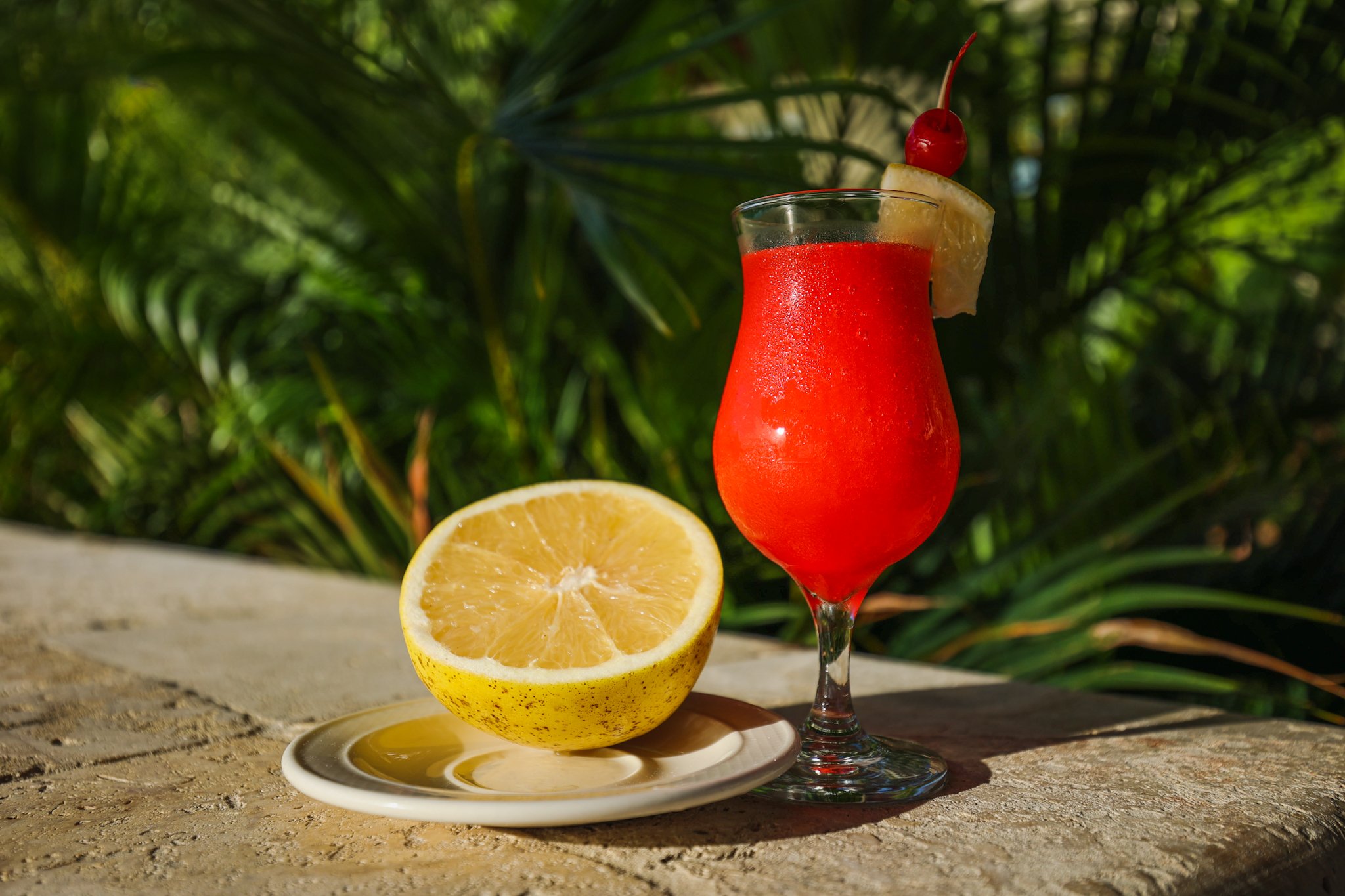 A vibrant red cocktail garnished with a cherry, lemon wedge, and lemon slice with a tropical greenery background.