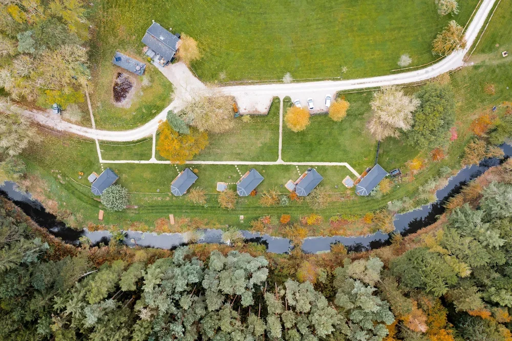 An aerial view of a residential area with six houses along a small river, surrounded by trees with fall foliage, and a grassy yard with a pathway and parked cars.