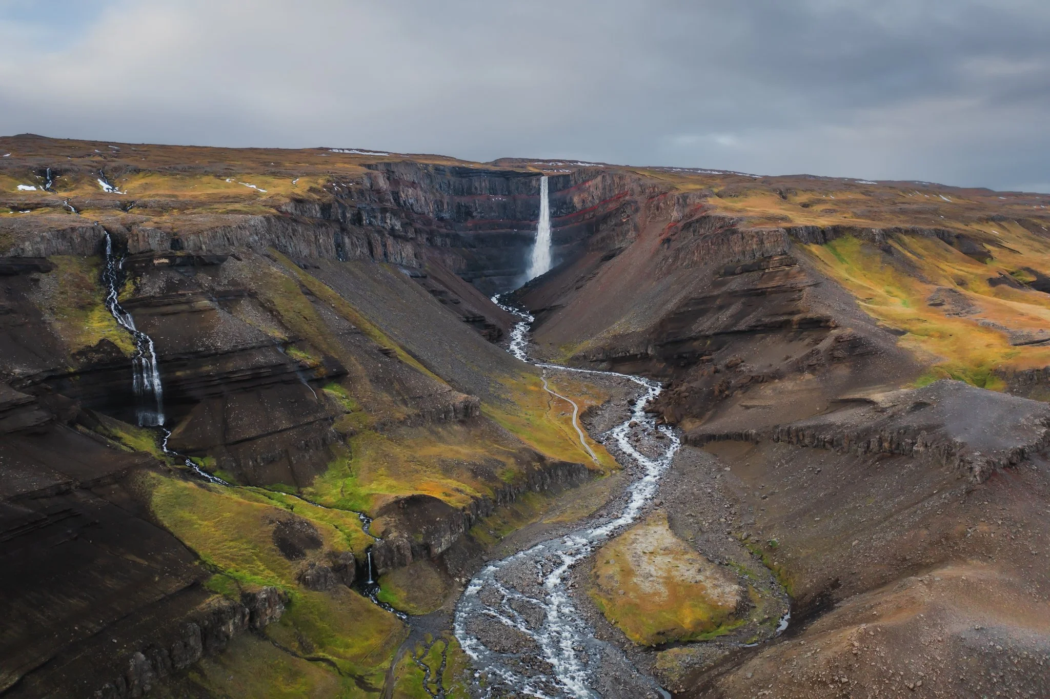 A landscape featuring a waterfall flowing down a cliff, with a stream running through a valley surrounding volcanic terrain and patches of grass and snow.