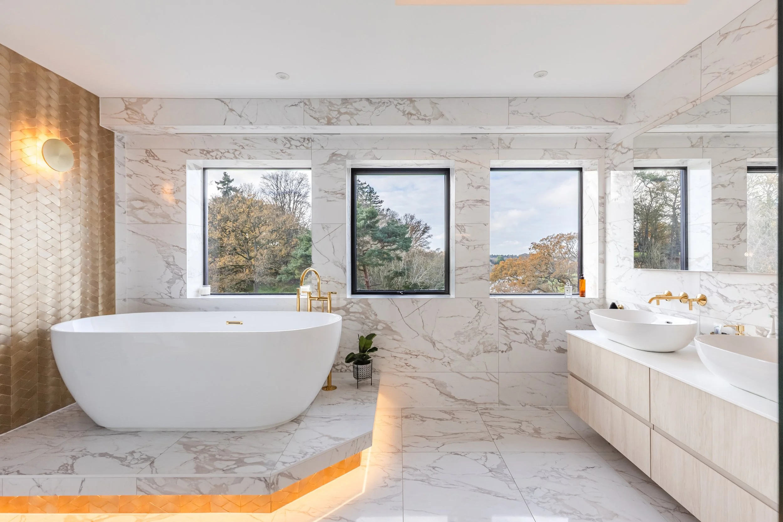 A luxurious bathroom featuring a white freestanding bathtub with gold fixtures, a marble countertop with vessel sinks, large windows showing trees outside, and elegant marble and gold decor accents.