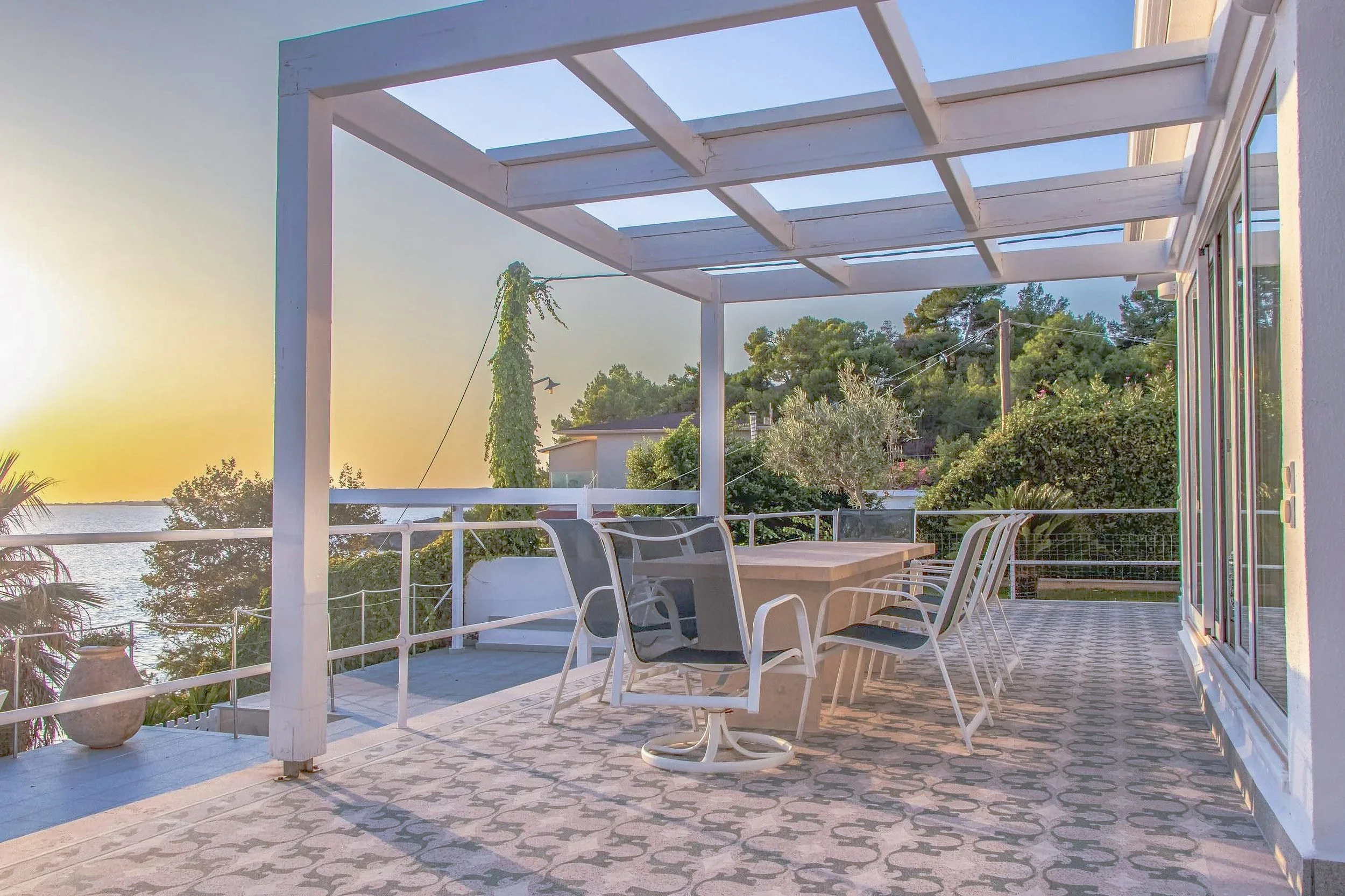 A backyard patio with a patterned floor, white railing, outdoor dining table with chairs, and a pergola with no cover, overlooking trees, bushes, and a body of water in the distance during sunset.