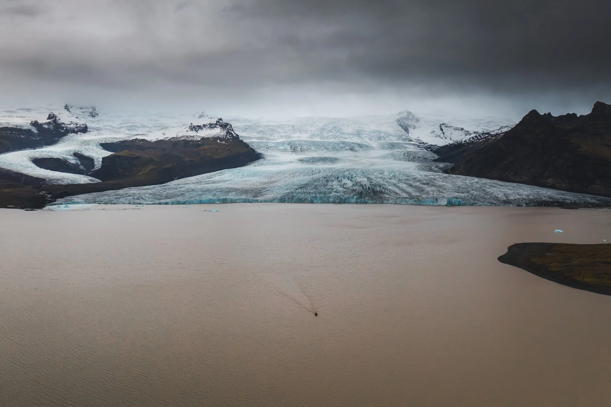 A landscape photo of a glacier flowing into a body of water, surrounded by dark rocky mountains, under a cloudy sky.