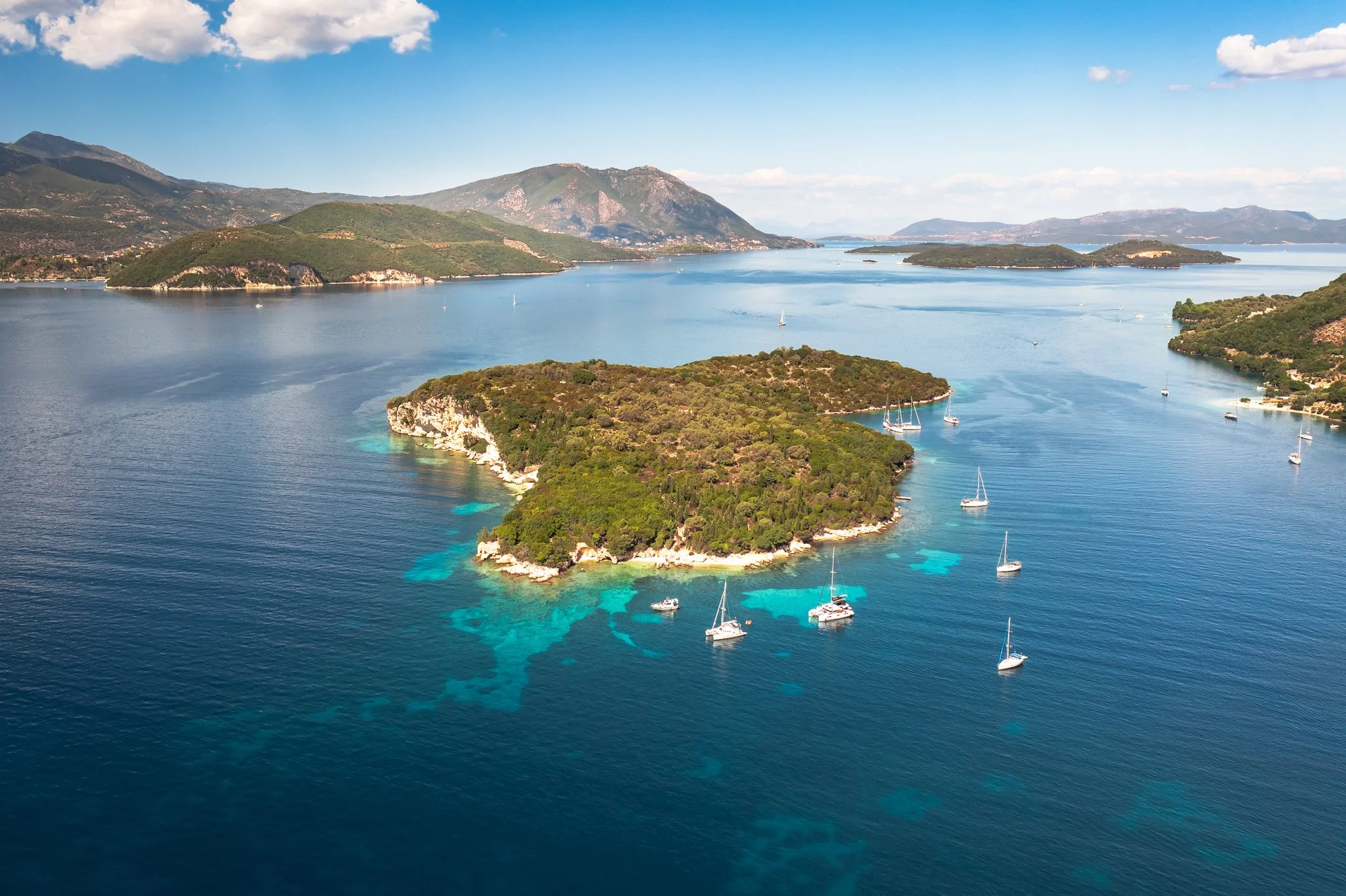 Aerial view of a coastal landscape with green islands and sailboats on clear blue water, mountains in the background, and a partly cloudy sky.
