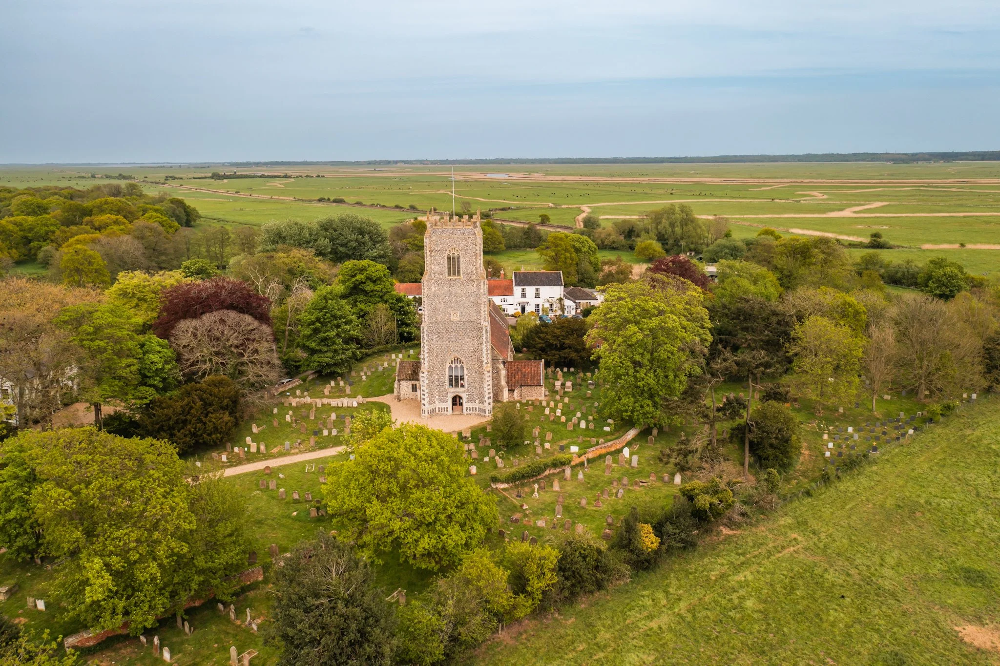 Aerial view of a church with a graveyard, surrounded by green trees, with open fields and farmland extending into the horizon under a cloudy sky.