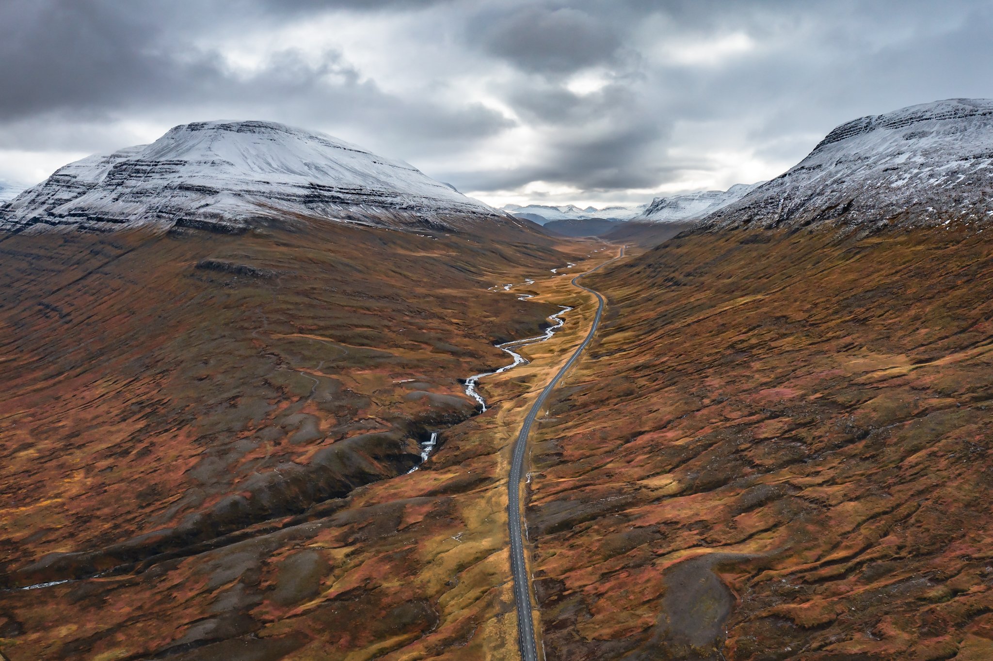 A scenic view of snow-capped mountains over a valley with a winding road and a small river, under a cloudy sky.