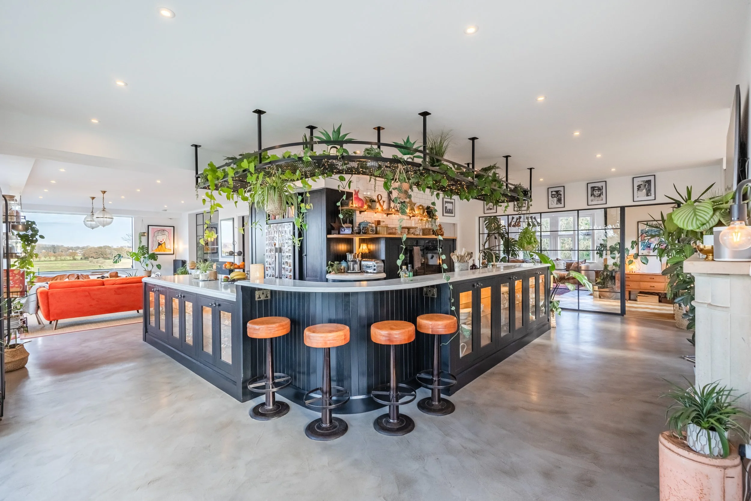 Interior of a modern open-concept living space with a black kitchen island topped with plants and decorative string lights, surrounded by barstools, a coral sofa, and framed photos on the walls.