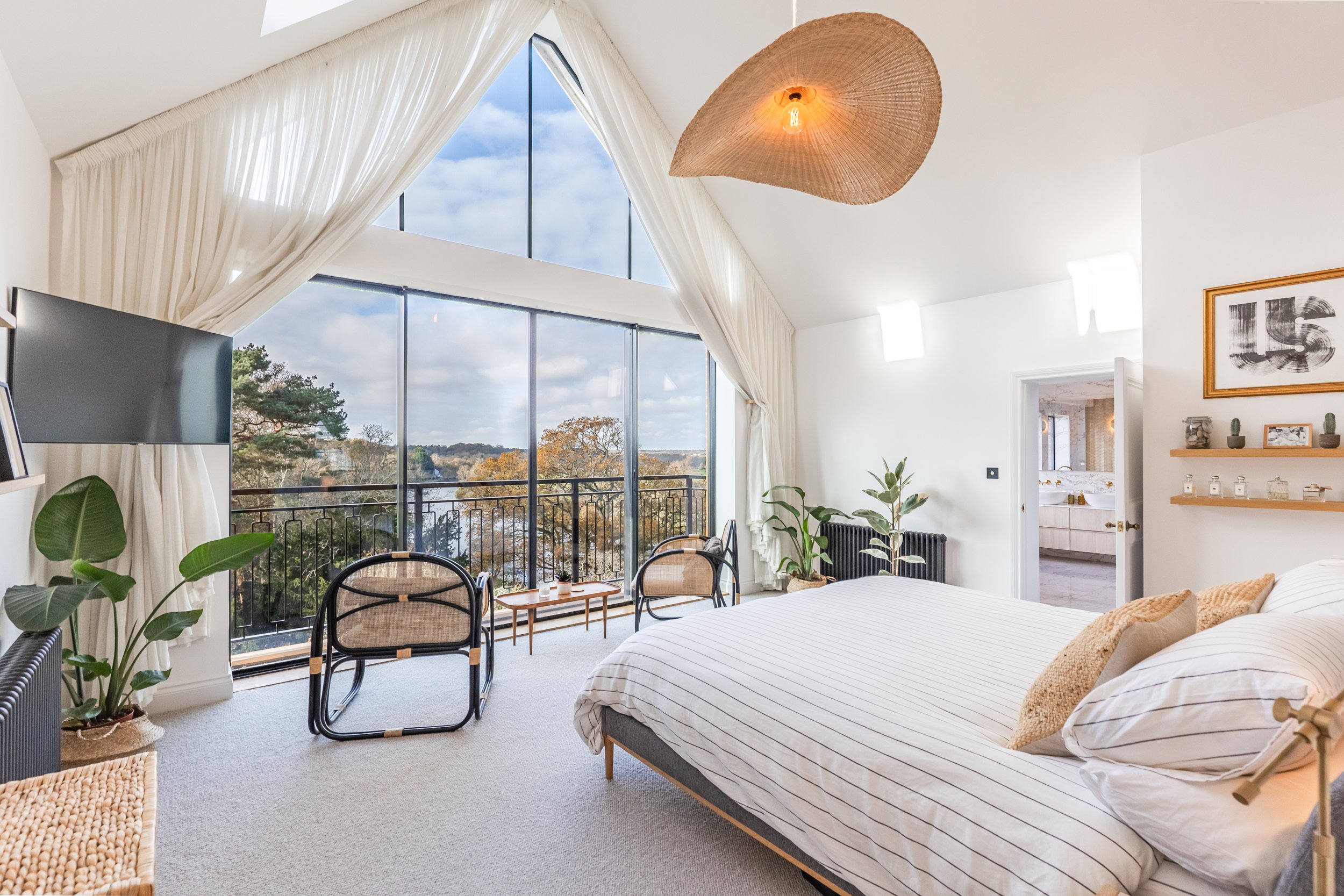 Bright bedroom with large triangular window, white walls, beige carpet, and white bedding with beige pillows. A black flat-screen TV is visible, along with potted plants, a woven chair, and a hanging wicker light fixture. There is a balcony with blac