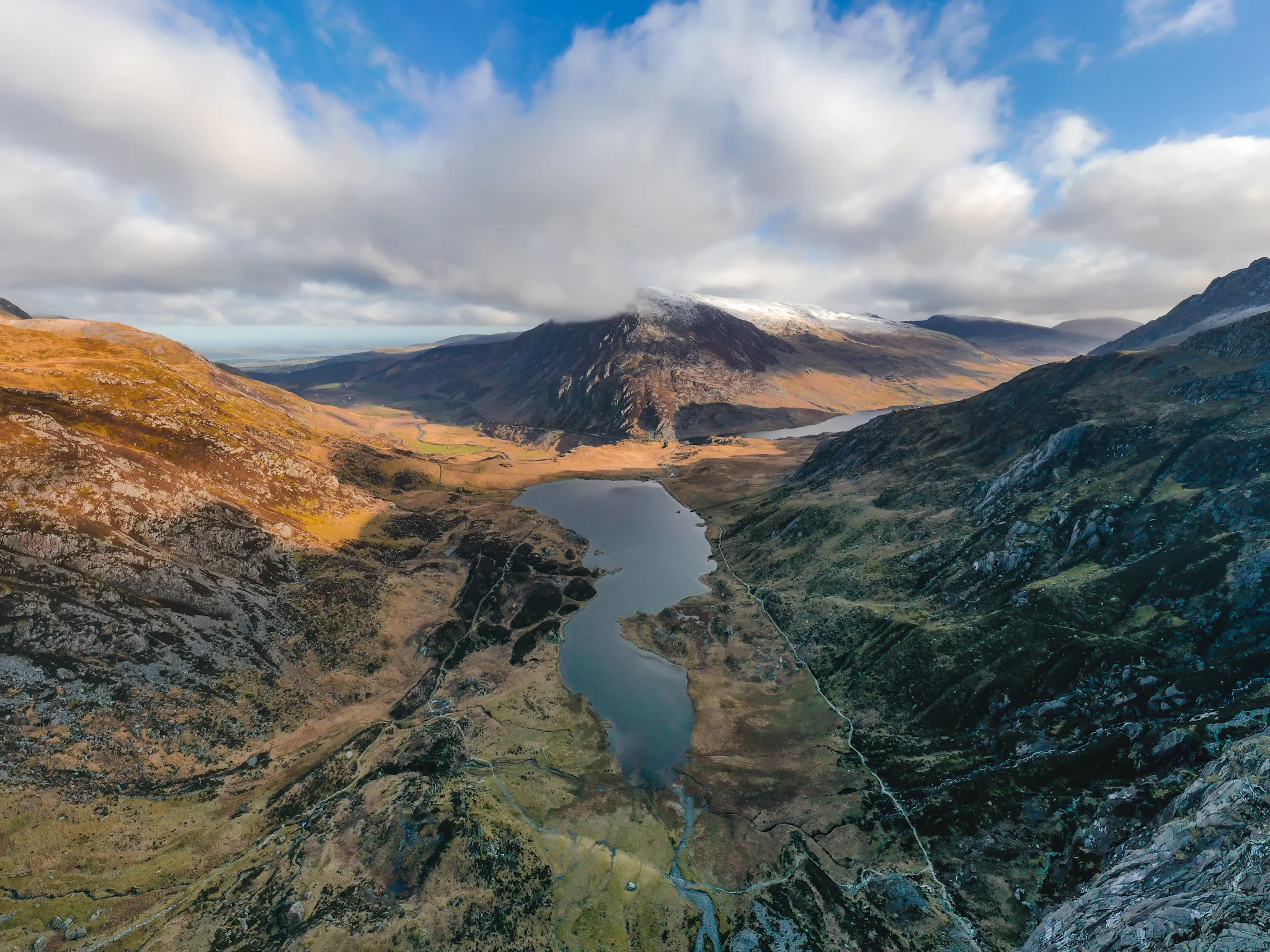 A scenic landscape of a mountain valley with a winding river, surrounded by rugged hills and mountains with some snow on the peaks, under a partly cloudy sky.