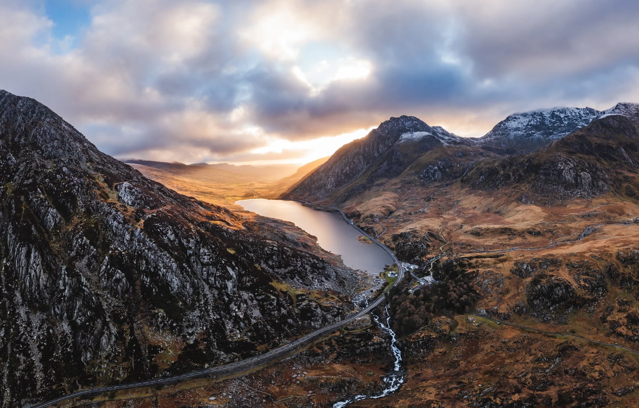 Aerial view of a mountainous landscape with a river running through the valley, surrounded by rocky peaks and patches of snow, with a cloudy sky at sunset.
