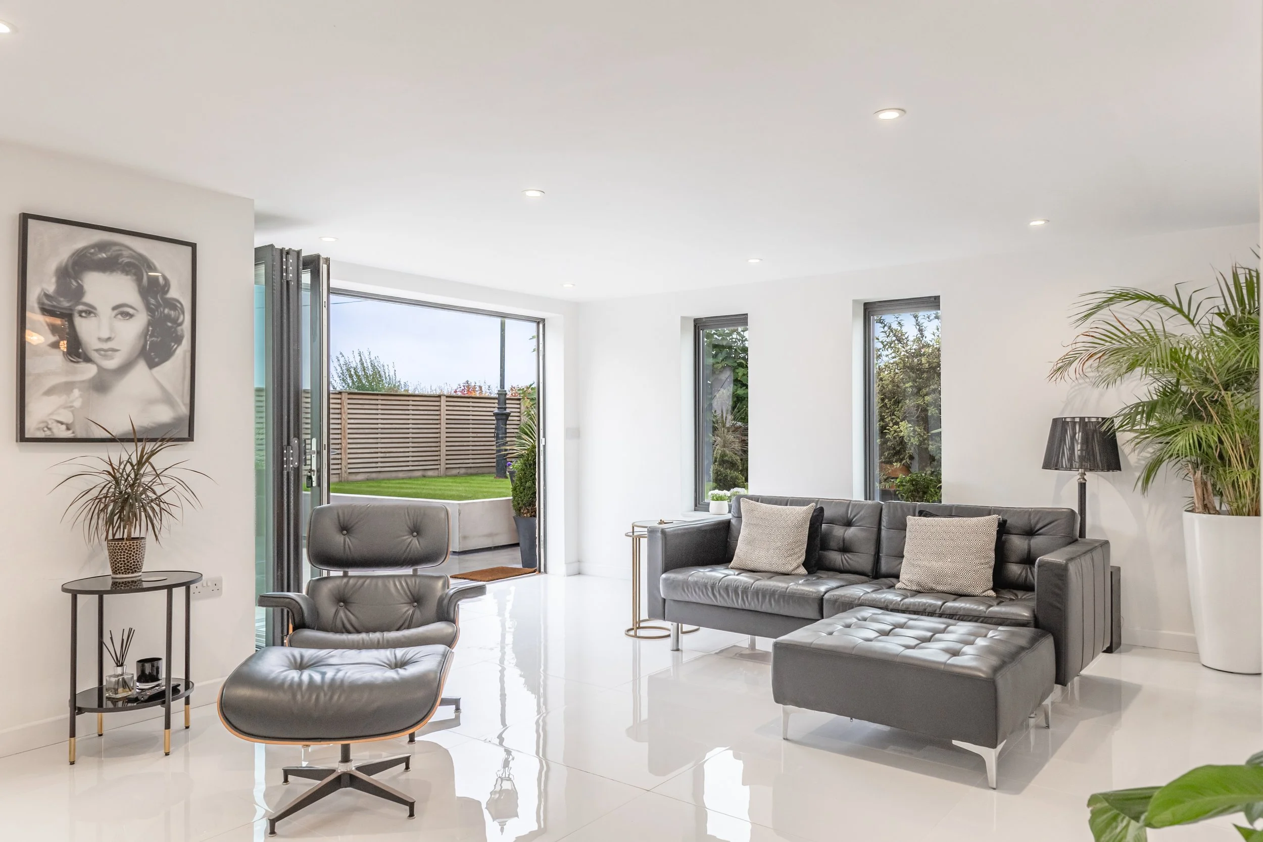 Modern living room with white walls, large windows, gray furniture, black and white portrait on the wall, plants, and a black table with decor.