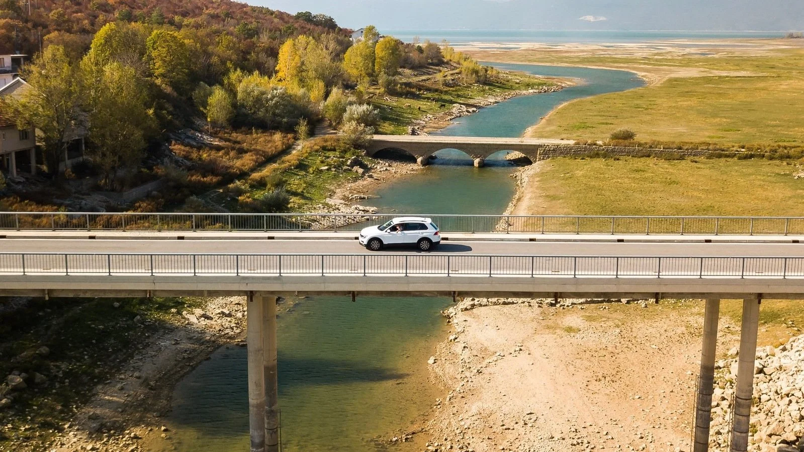 A white SUV driving across a bridge over a river in a rural area, with fields, trees, and a distant shoreline under a clear blue sky.