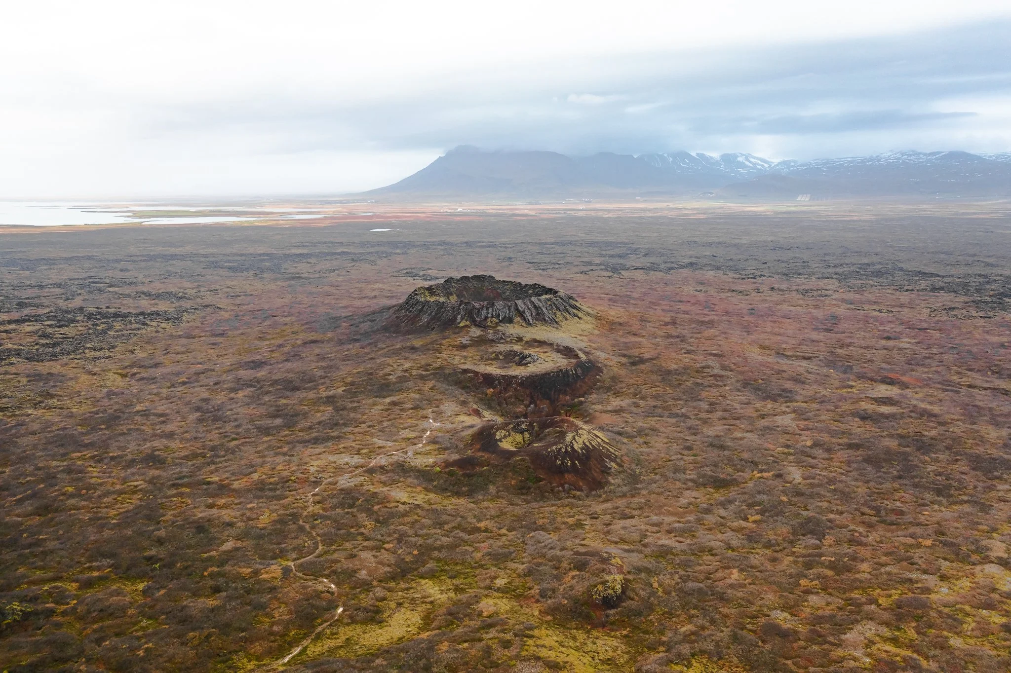 A wide landscape with volcanic terrain, moss-covered ground, a small volcanic cone in the foreground, and mountains with snow on their peaks in the background under a cloudy sky.