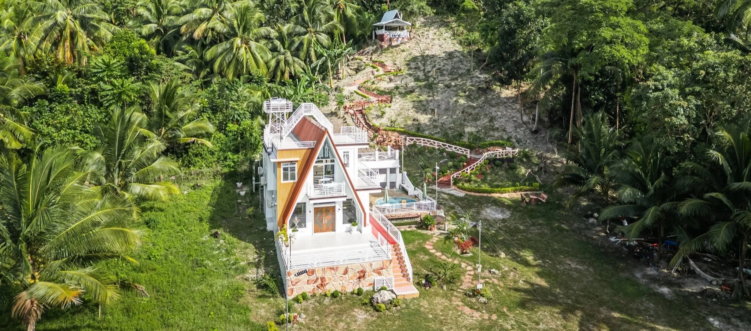 A house with A-frame design surrounded by lush greenery, tropical trees, and a garden with pathways and outdoor seating, situated on a hillside with stairs leading up to the house.