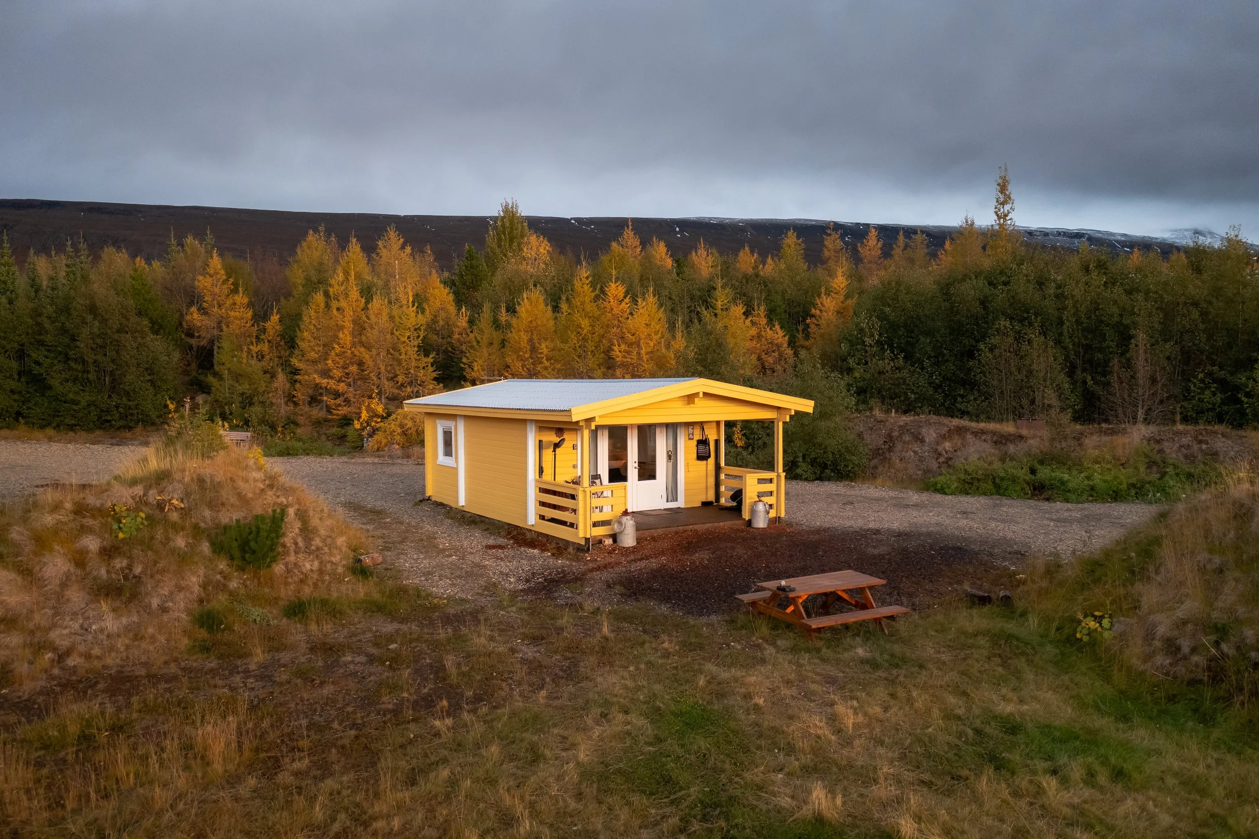 Yellow tiny house with a porch, located outdoors in a natural setting with trees and mountains in the background under a cloudy sky.