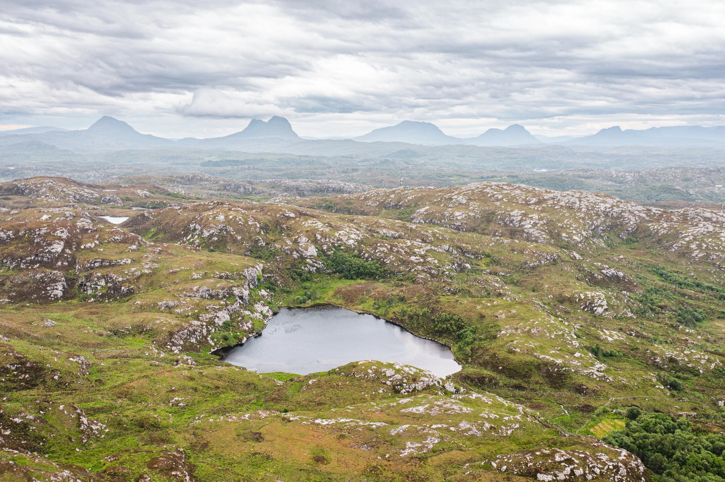 A mountainous landscape with rocky hills and a small lake in the foreground, with distant mountains underneath a cloudy sky.